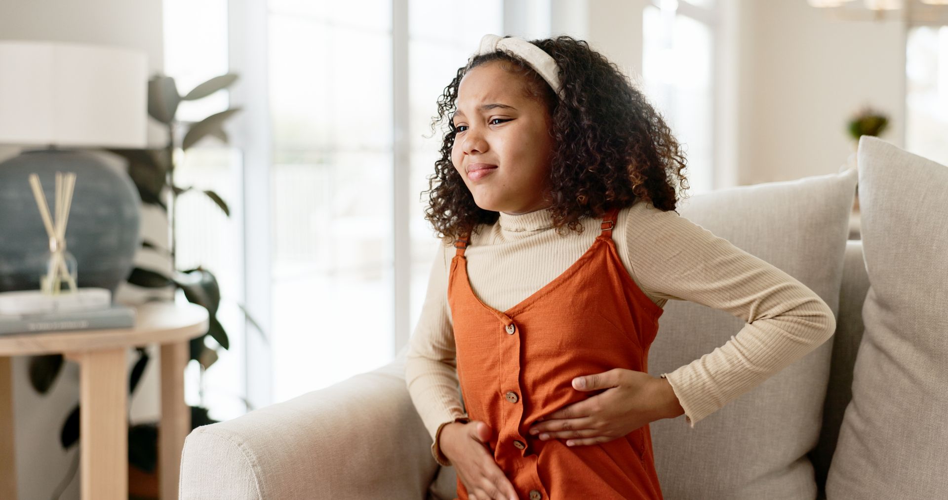 Une jeune fille en robe orange se tient le ventre, visiblement souffrante, assise sur un canapé à l'intérieur.