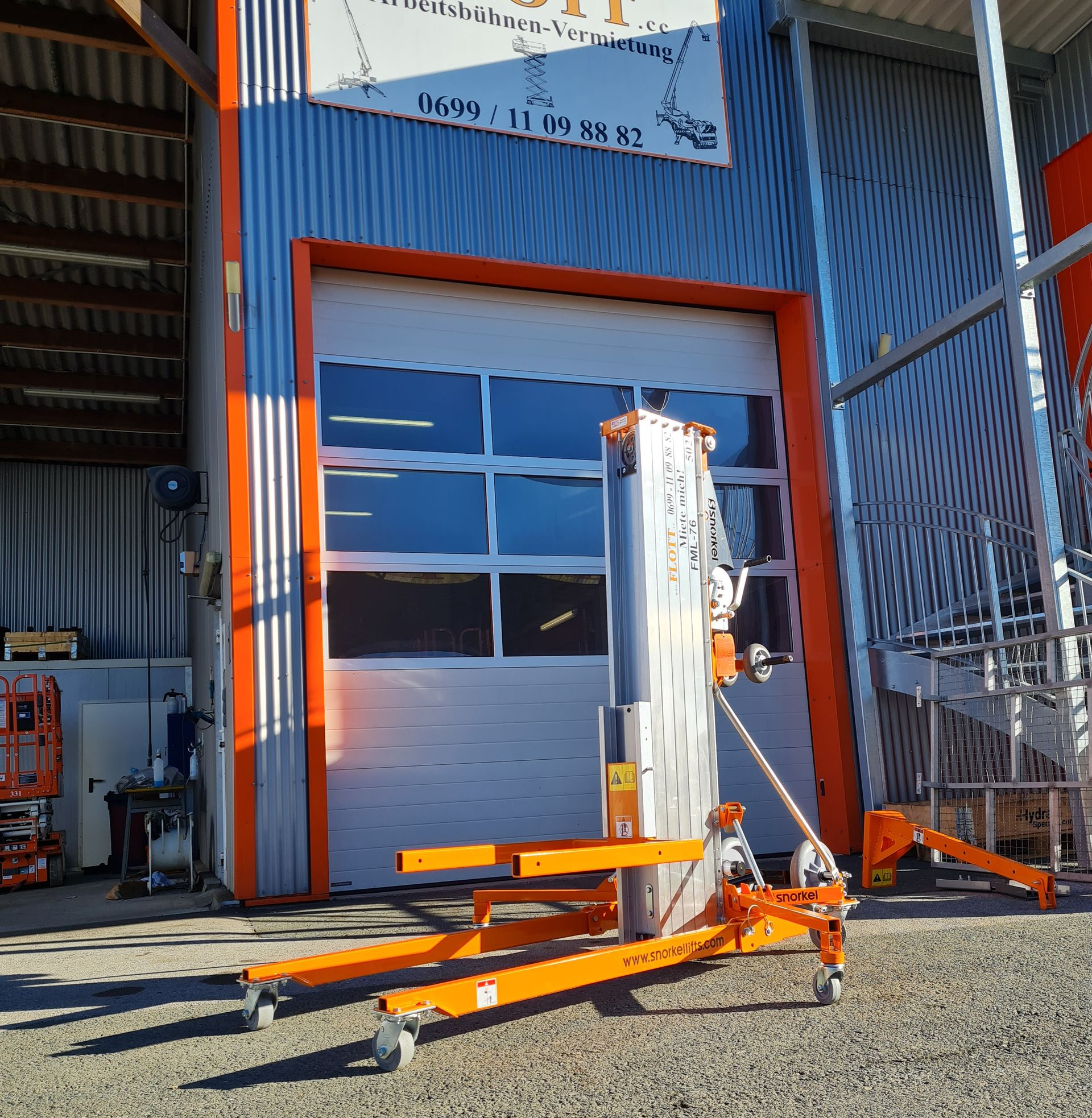 Vor einem Gebäude mit weißer Tür steht eine orangefarbene Aufzugsanlage.