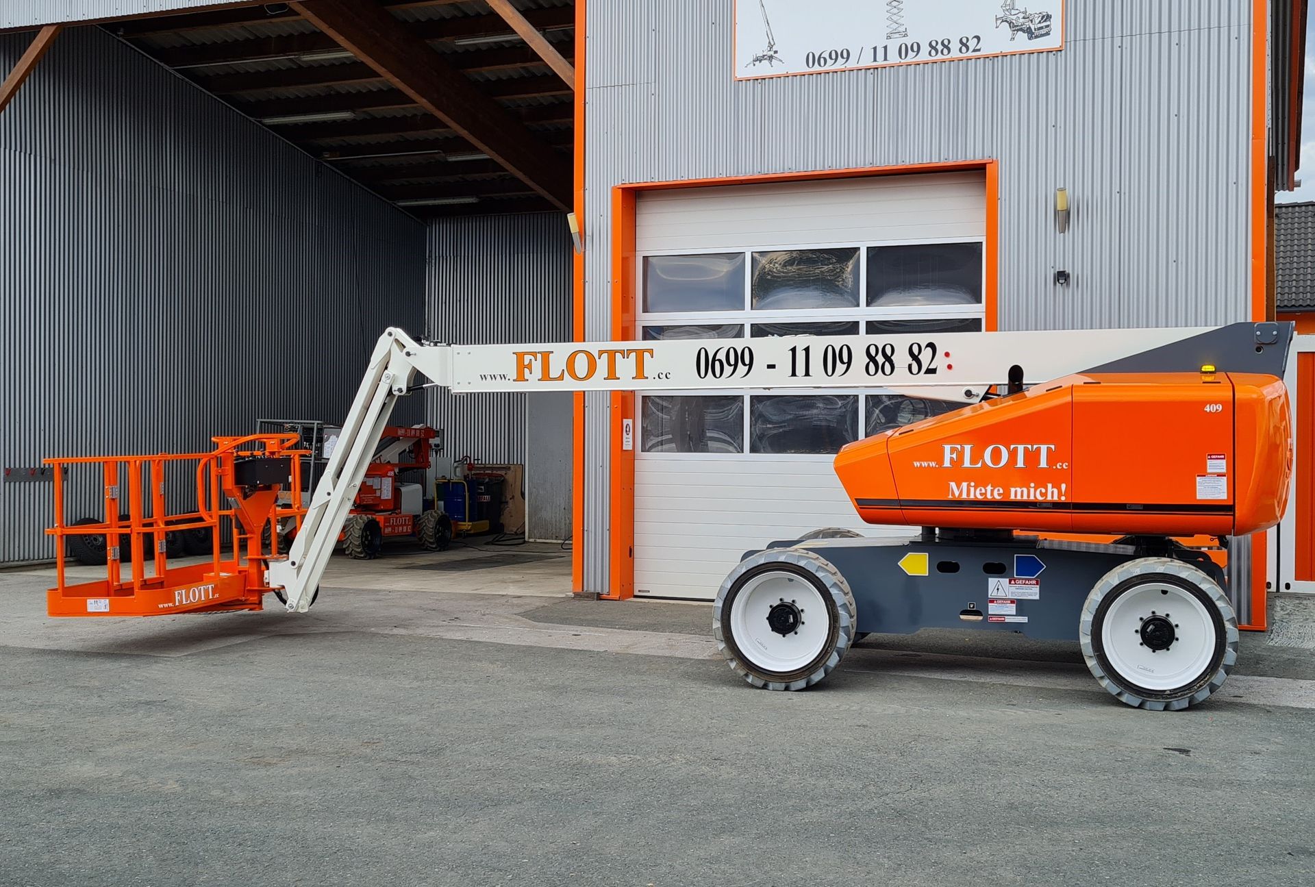 Eine orangefarbene Hubarbeitsbühne der Marke „FLOTT“ parkt vor einem Gebäude mit weißer Tür.