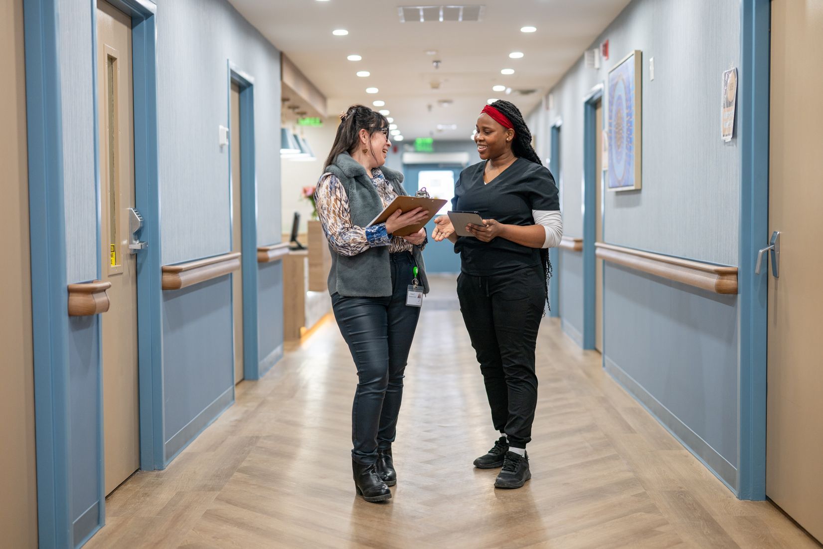 Two women are standing in a hospital hallway looking at a clipboard.