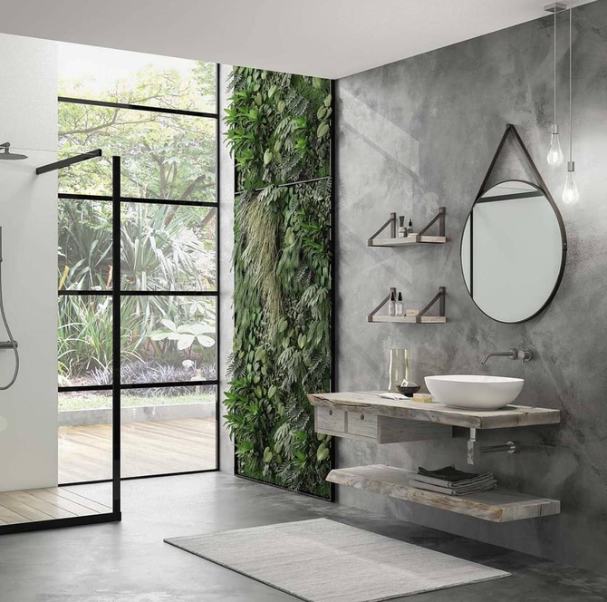 Bagno moderno con pareti in cemento, lavabo sospeso, specchio rotondo e doccia in vetro accanto a una parete verde con piante.