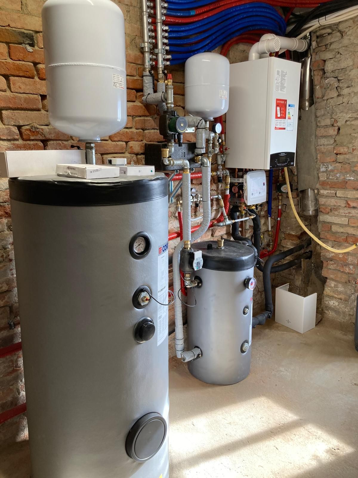 Locale tecnico nel seminterrato con scaldabagni, serbatoi, tubature e caldaia addossati a pareti in mattoni a vista.