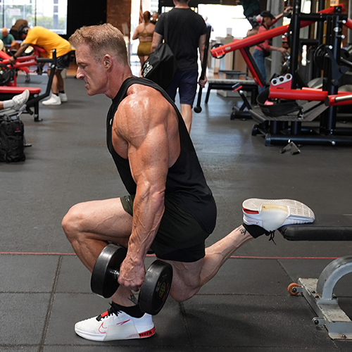 Ron Matthews is squatting down while holding a dumbbell in a gym.