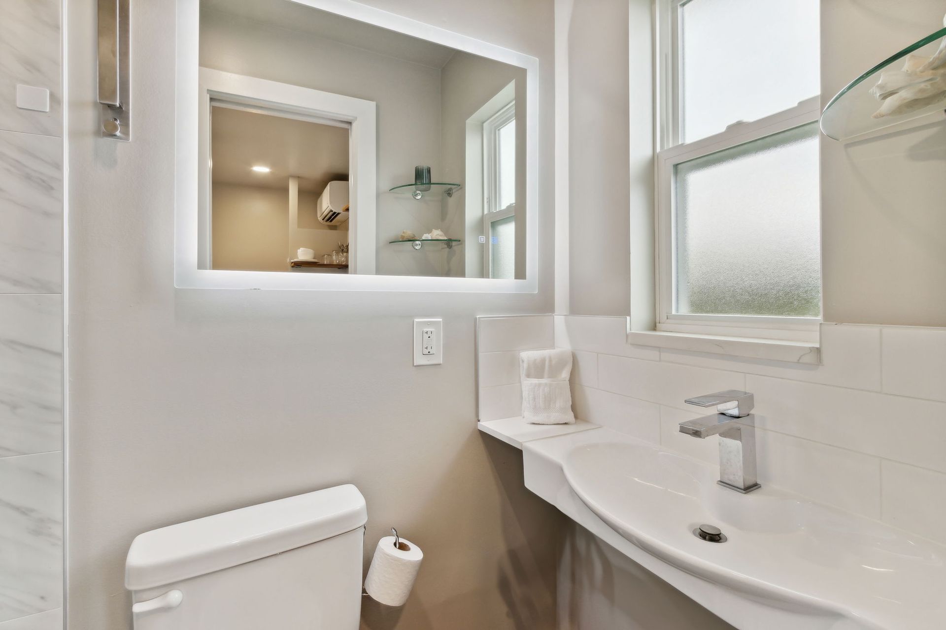 A bathroom with a toilet, sink, LED lit mirror and window.