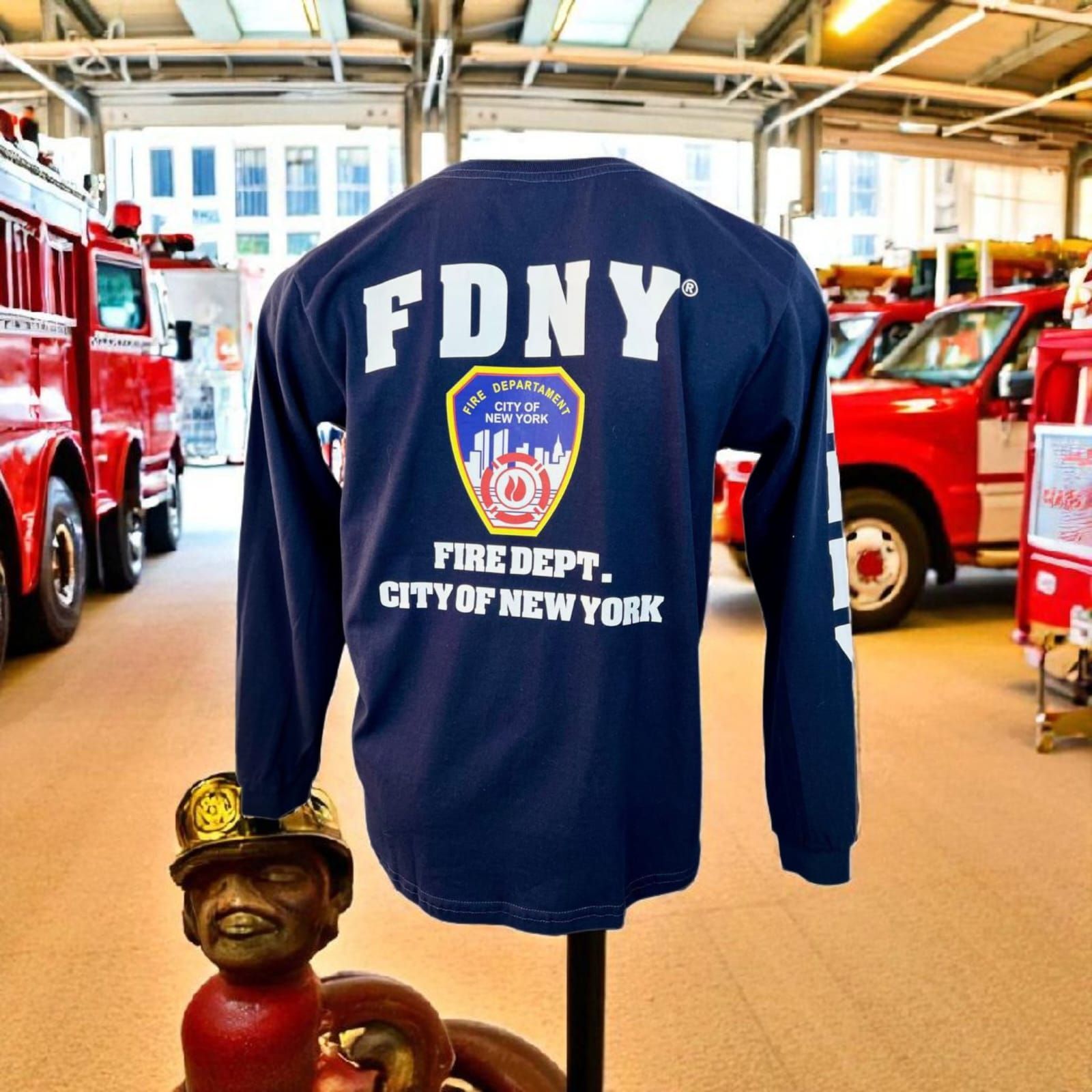 Camisa de manga larga de la Marina del FDNY expuesta en una estación de bomberos o museo, con camiones de bomberos al fondo.