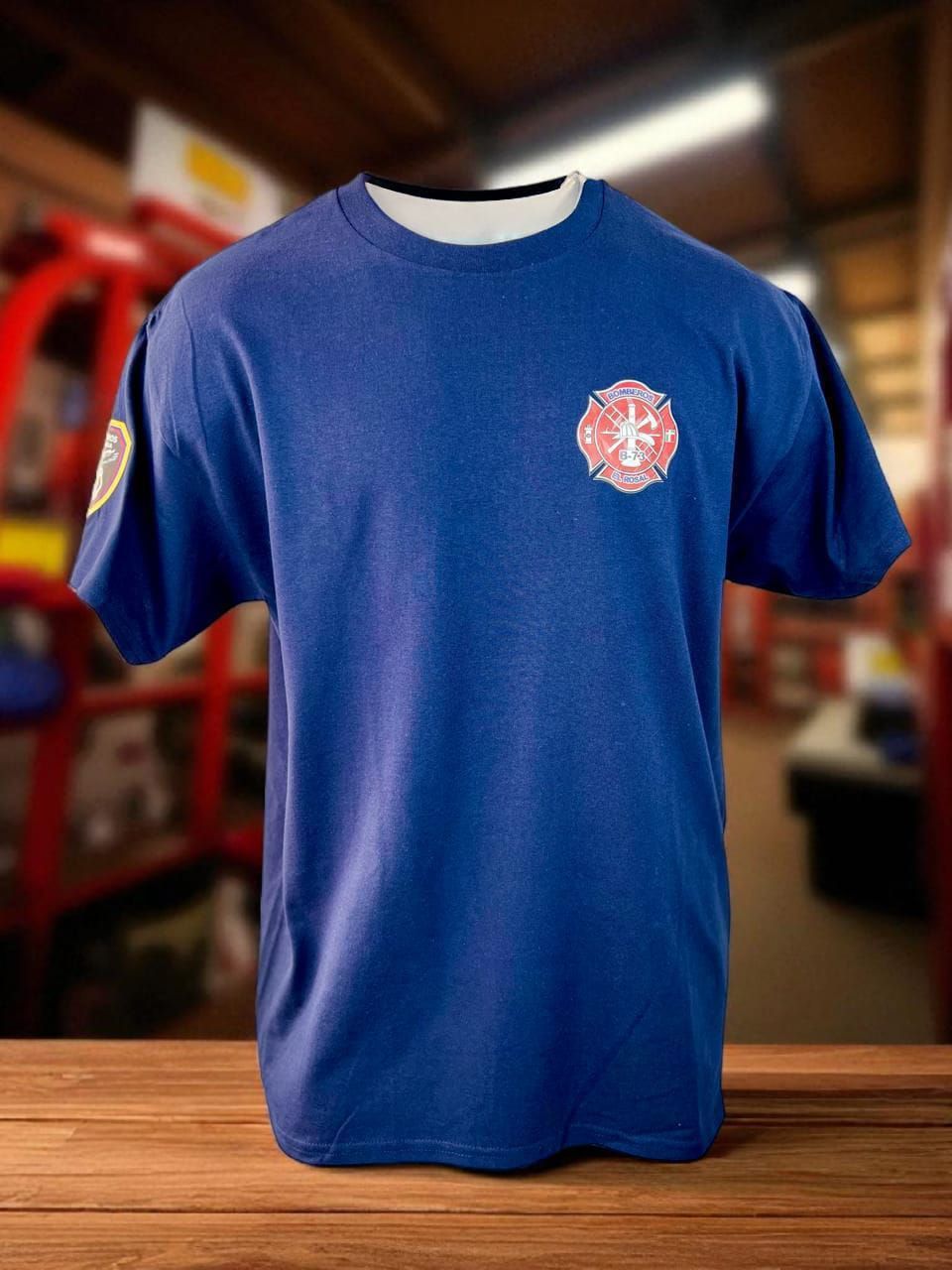 Camiseta azul de “Bomberos Colombia” expuesta sobre una mesa de madera, con un vehículo de emergencia borroso al fondo.