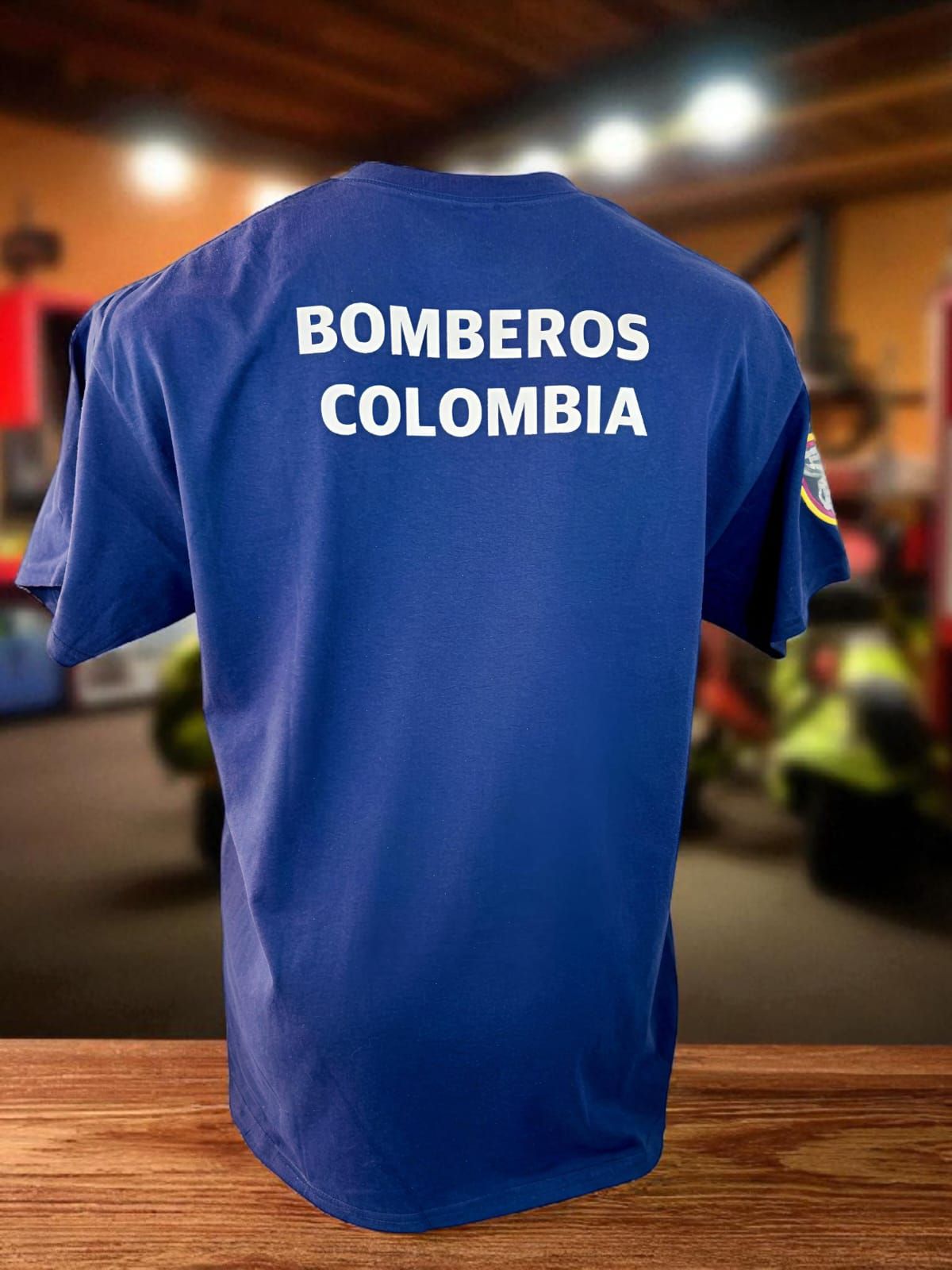 Camiseta azul de “Bomberos Colombia” expuesta sobre una mesa de madera, con un vehículo de emergencia borroso al fondo.