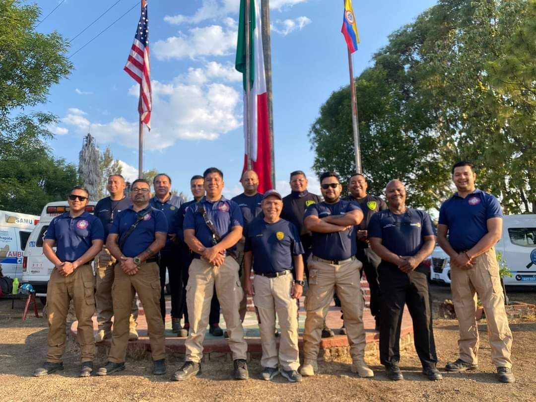 Grupo de personas con camisas azules y pantalones caqui posando al aire libre junto a banderas de Estados Unidos e Italia.