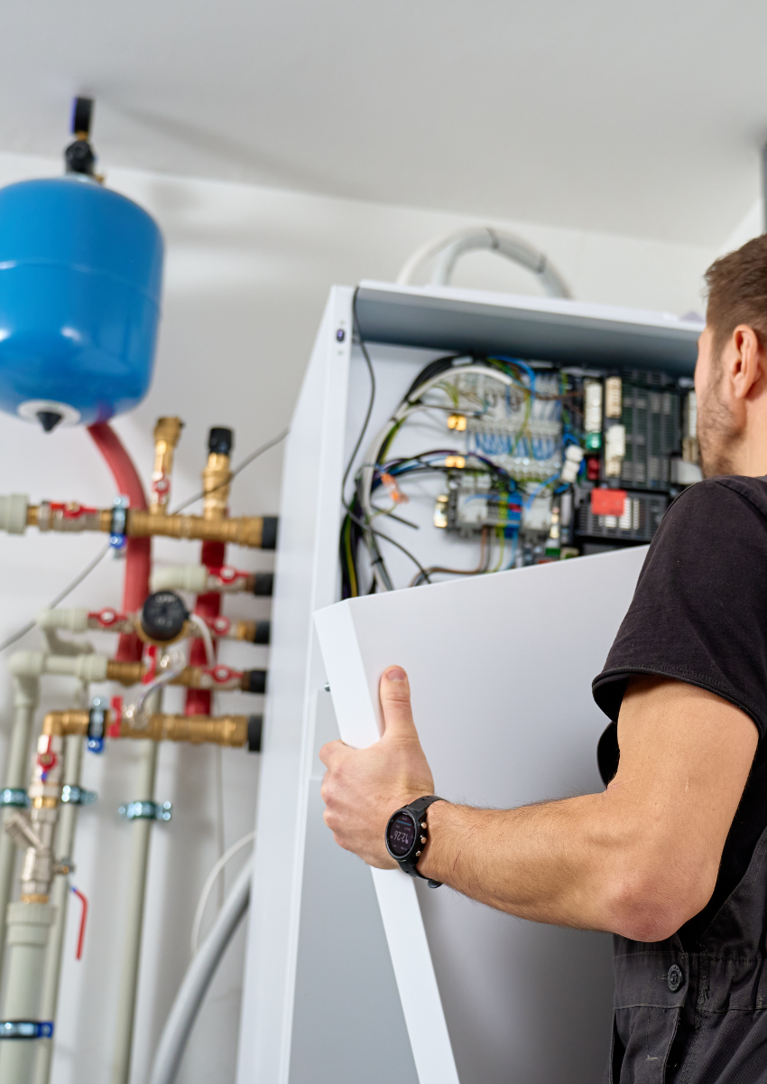 Technician carrying a white appliance beside an open boiler with exposed wiring and pipes