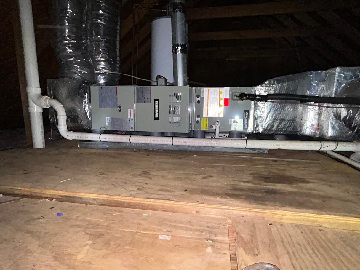 Attic HVAC unit with ductwork and pipes on wooden joists under a low roof