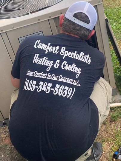 Worker kneeling by an outdoor HVAC unit, wearing a black Comfort Specialists Heating & Cooling shirt.