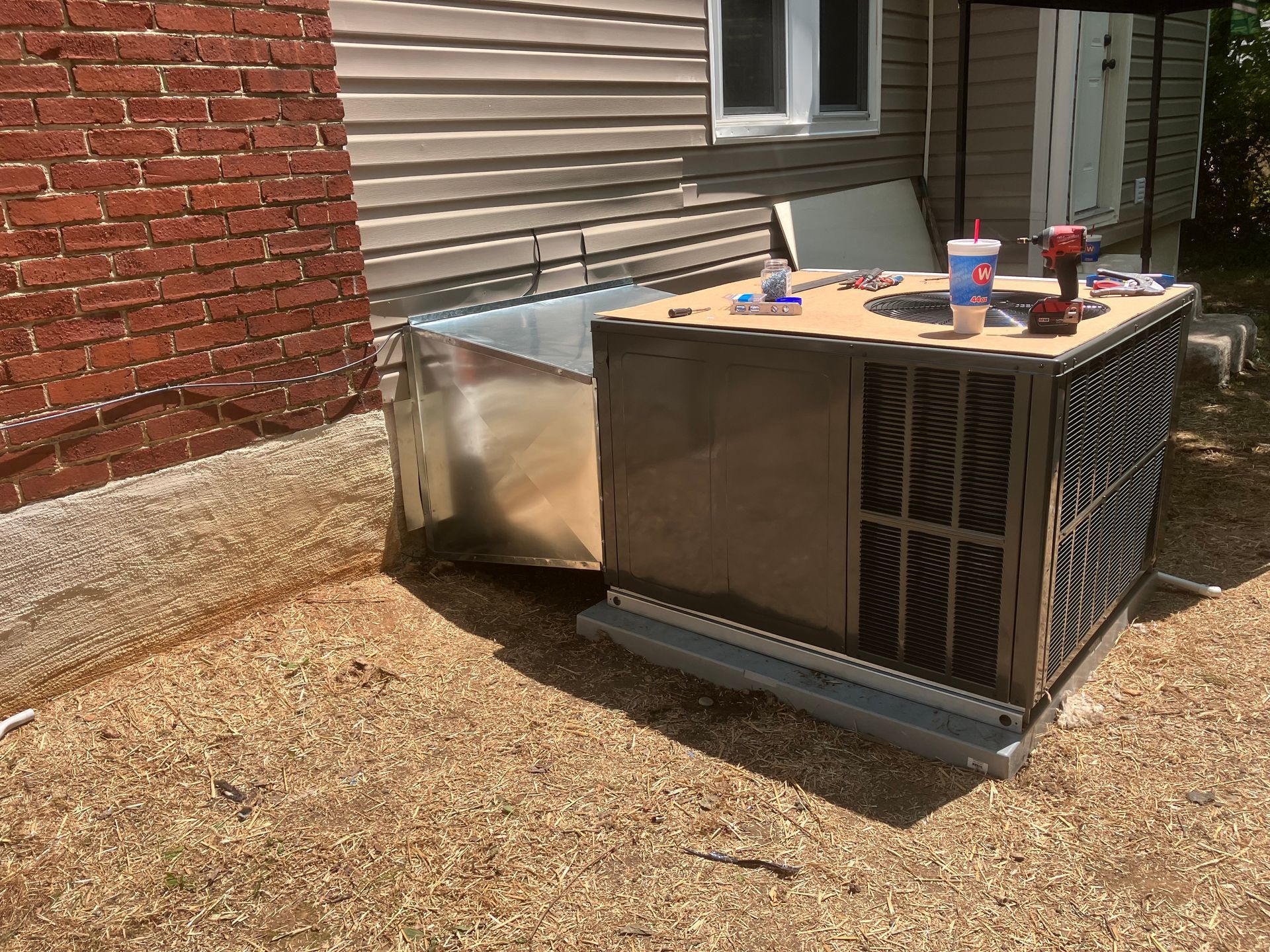 Outdoor HVAC unit beside a brick and siding house, with tools resting on top in a gravel yard.