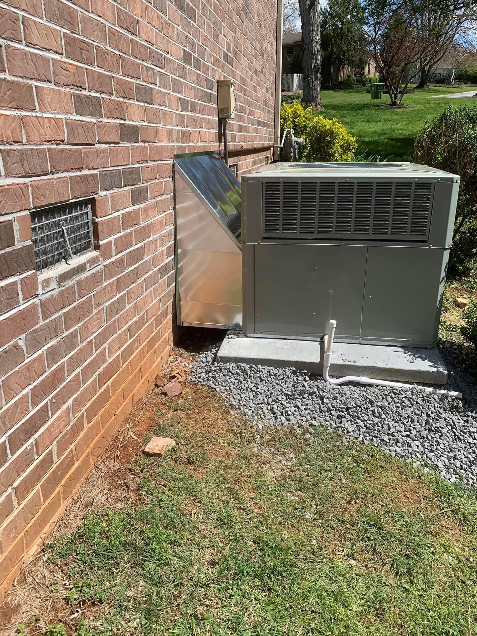 Outdoor HVAC unit beside a brick house wall on a gravel pad, with grass and shrubs nearby