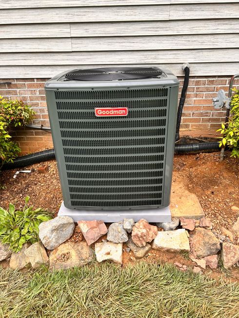 Outdoor air conditioner unit beside a house wall, with stones and mulch at the base.