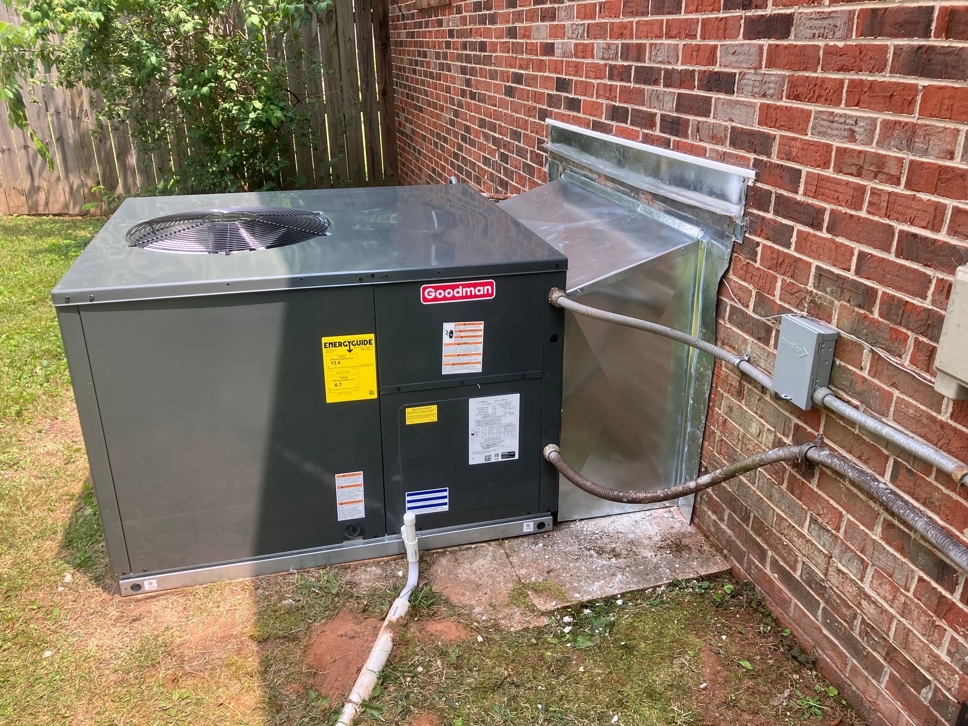 Outdoor HVAC unit beside a brick house with metal ductwork and electrical conduit attached