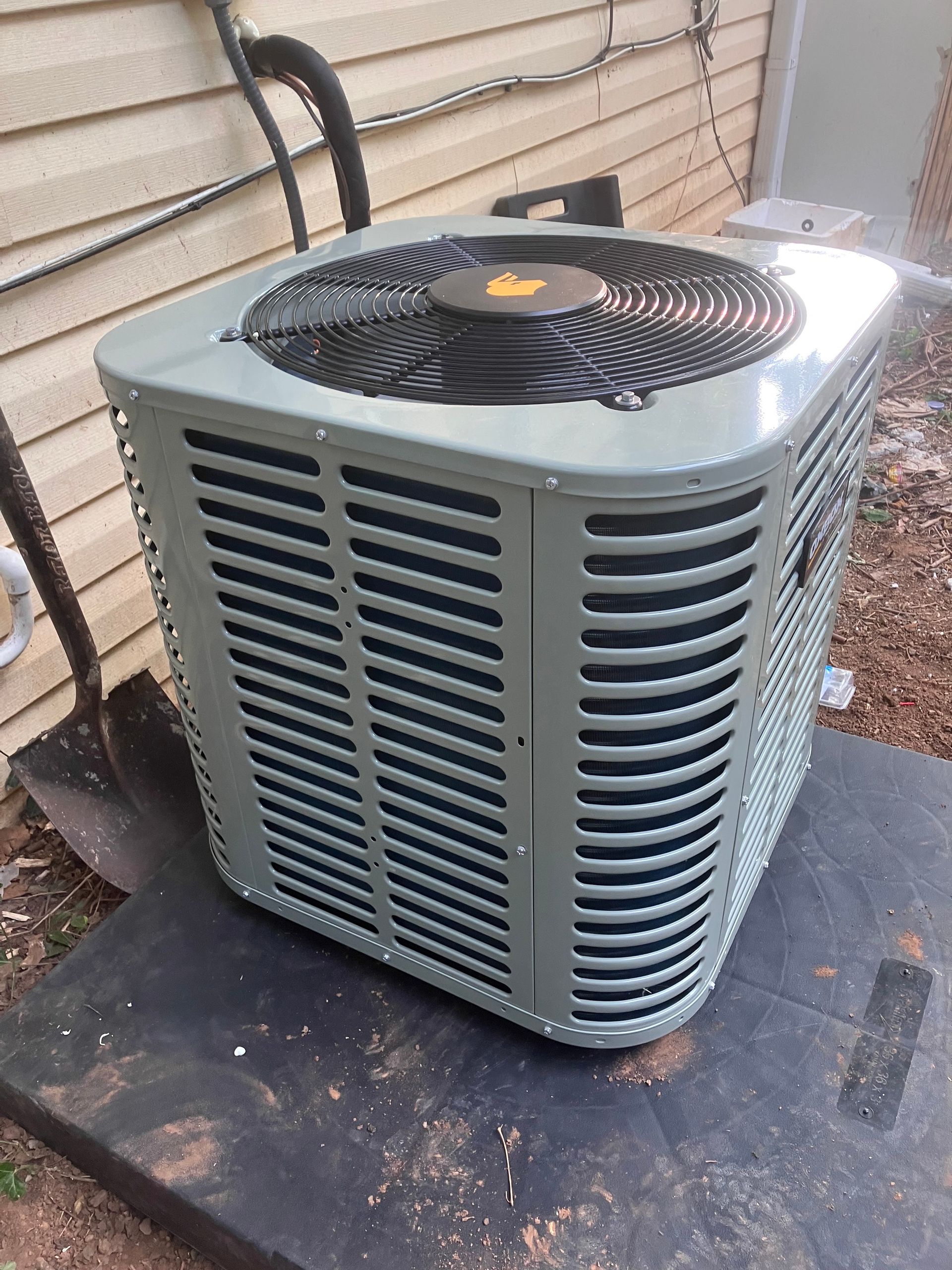 Outdoor central air conditioning unit beside a house wall and window, with grass and mulch nearby
