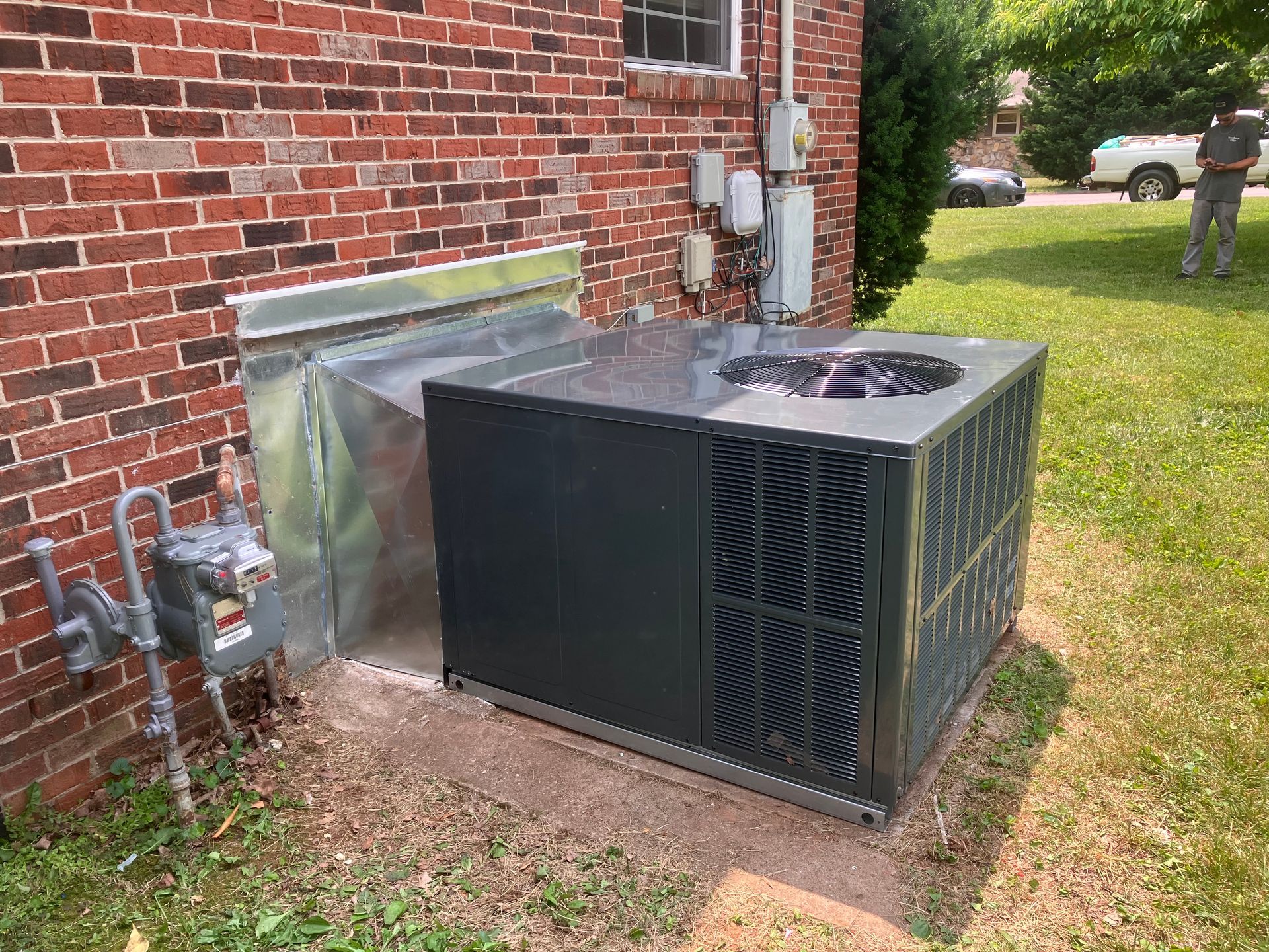 Outdoor HVAC unit beside a brick house, with metal ducting and utility connections.
