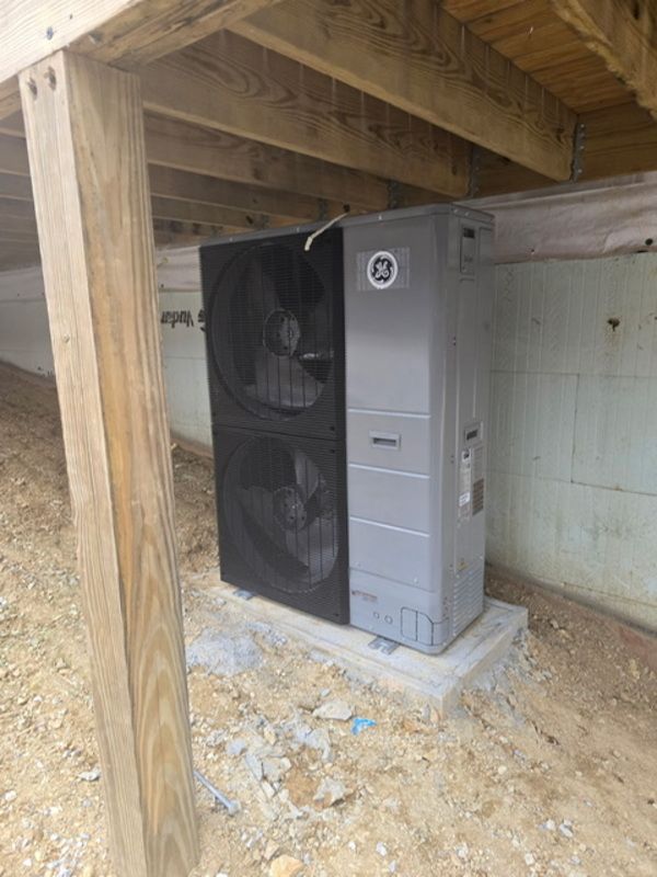 Outdoor HVAC unit under a deck beside a house wall, on a concrete pad.