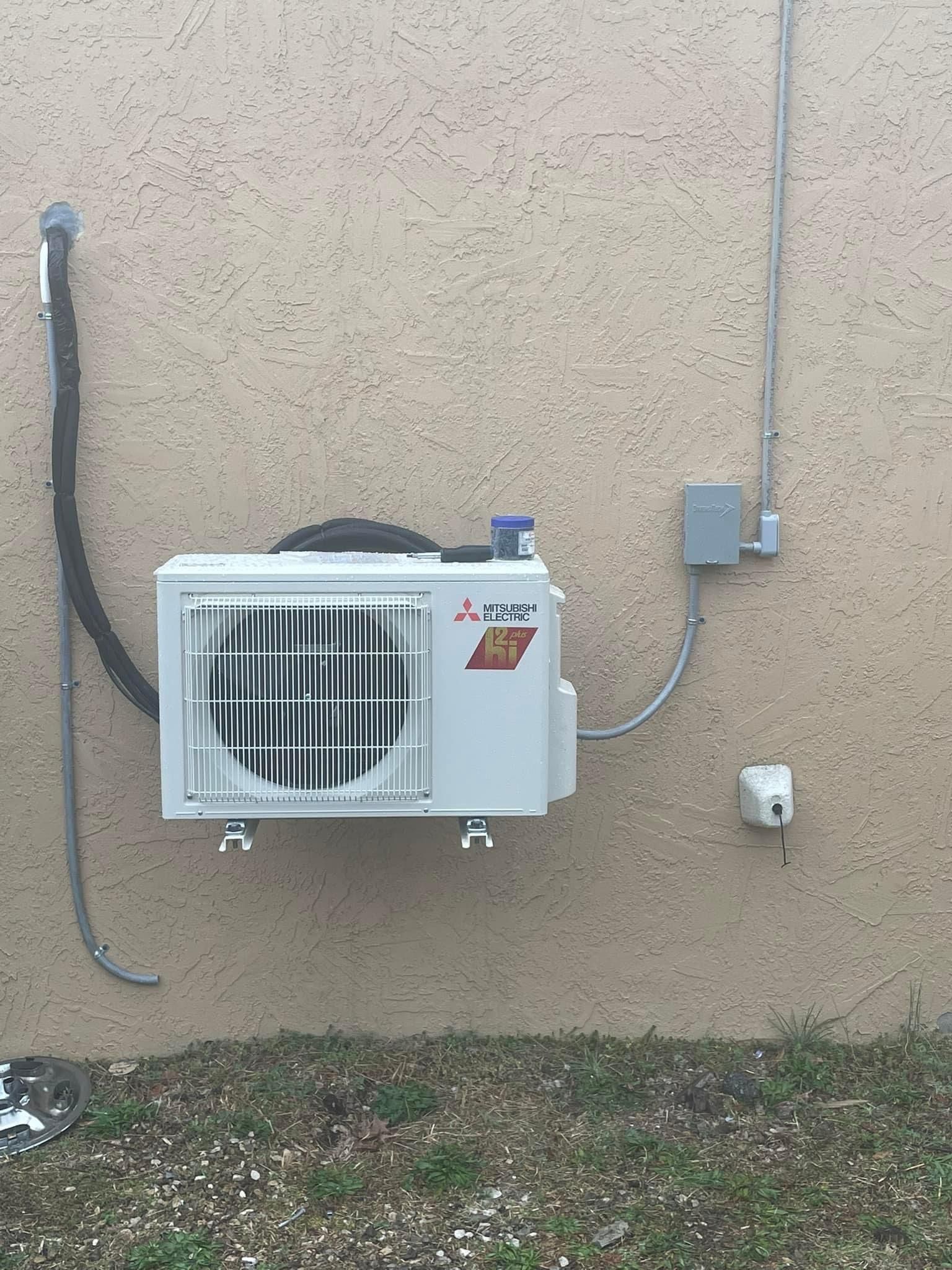 Exterior air conditioning unit mounted on a beige wall with visible conduit and electrical box.
