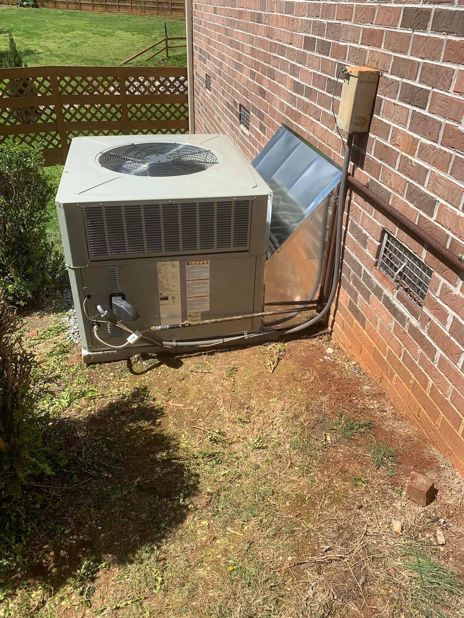 Outdoor air conditioning unit beside a brick house wall with a fan on top and a gray panel leaning against it.