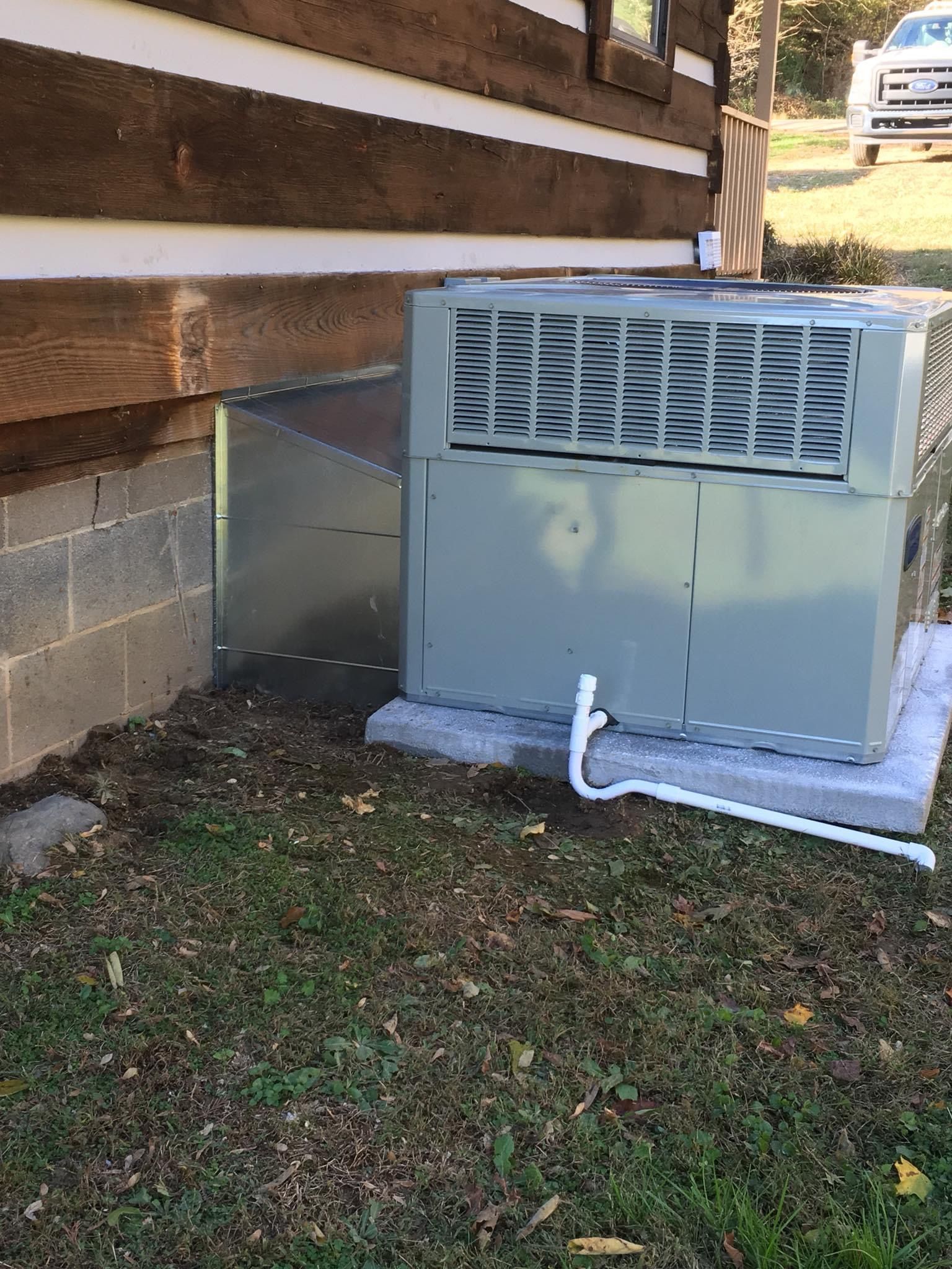 Outdoor HVAC unit beside a house, with a white pipe and cinder block foundation, on a grassy yard.