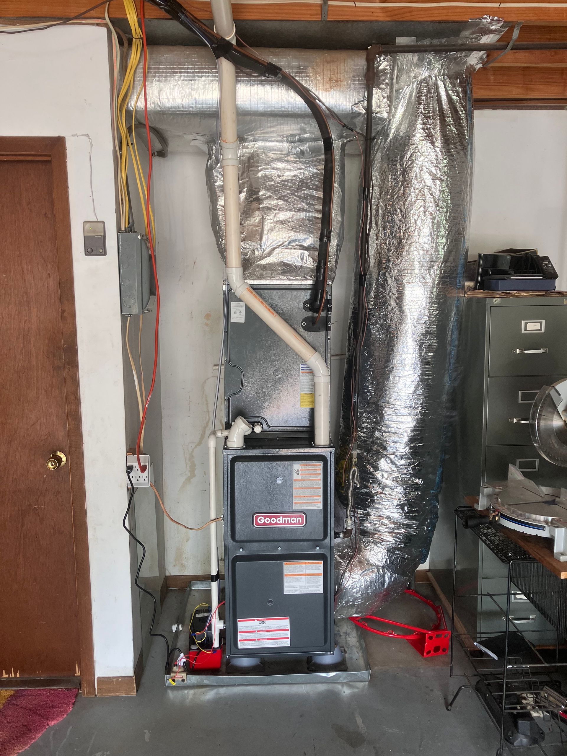 Basement furnace and HVAC ductwork beside a door, with exposed pipes and foil insulation.
