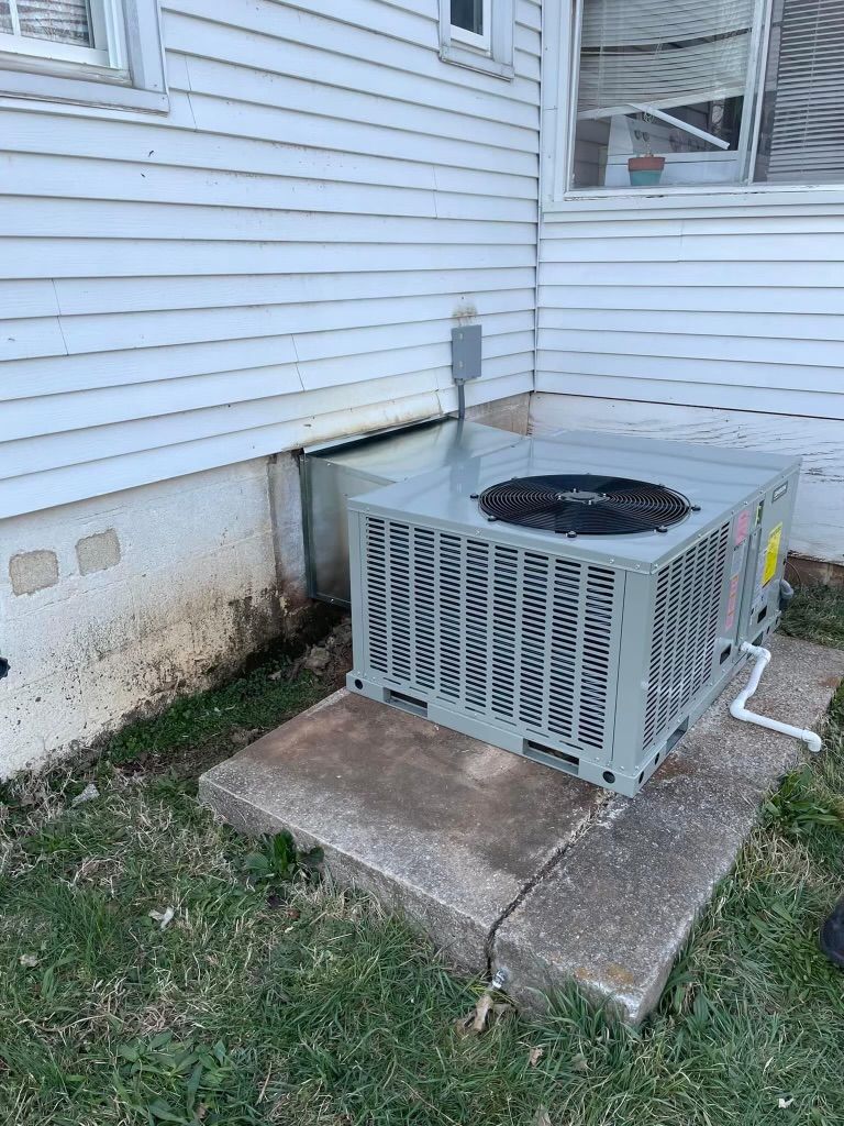 Outdoor air conditioner unit beside a white house, sitting on a concrete pad near the wall.
