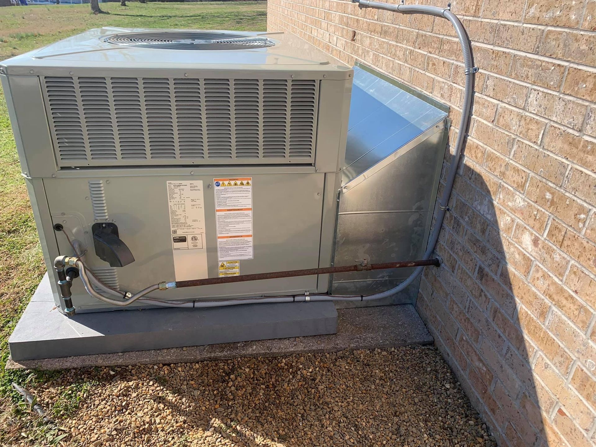 Outdoor HVAC unit beside a brick wall, with visible conduit and pipes on a concrete pad.