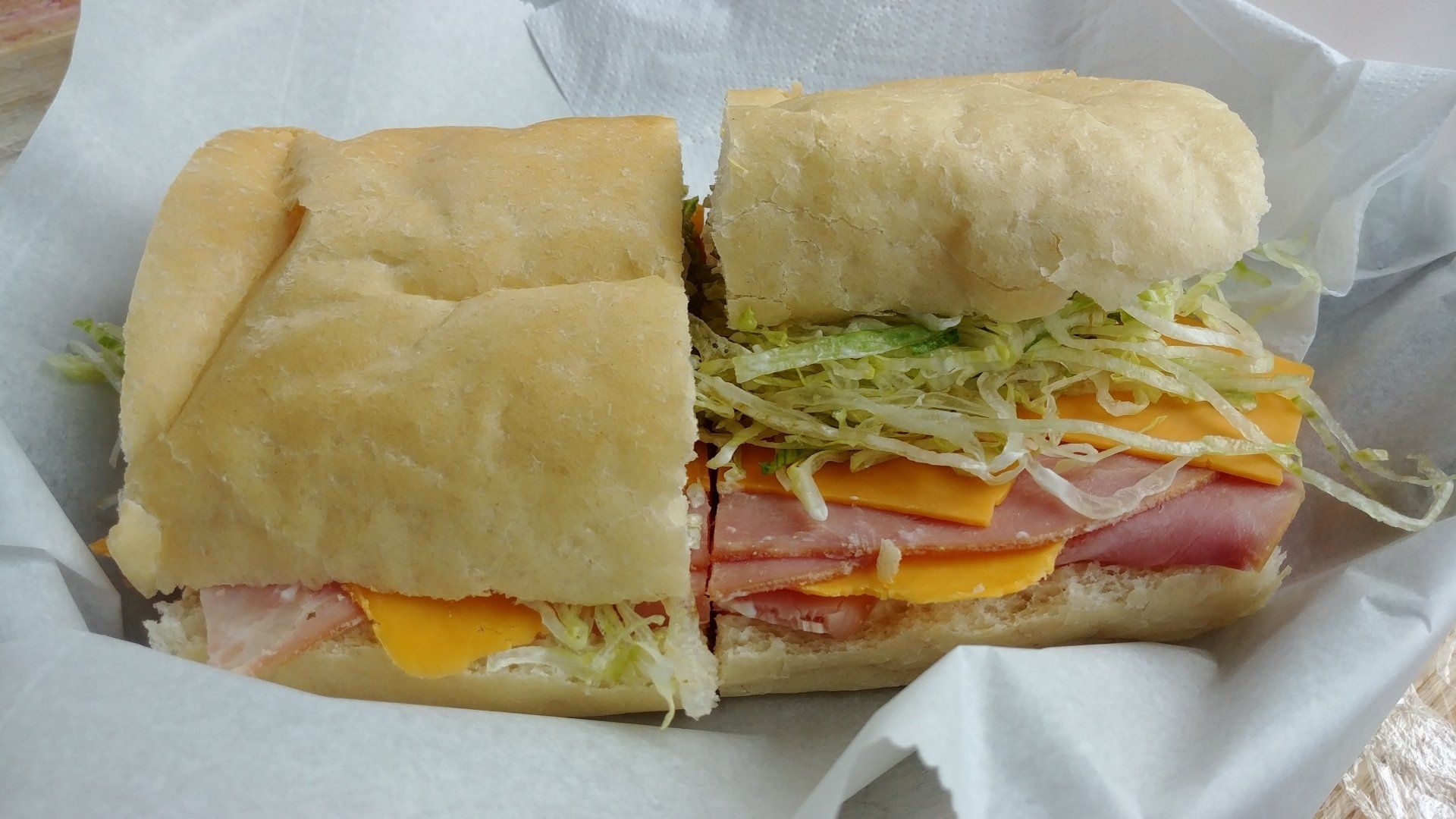 A ham and cheddar cheese sub sandwich topped with shredded lettuce, served on white deli paper in a basket.