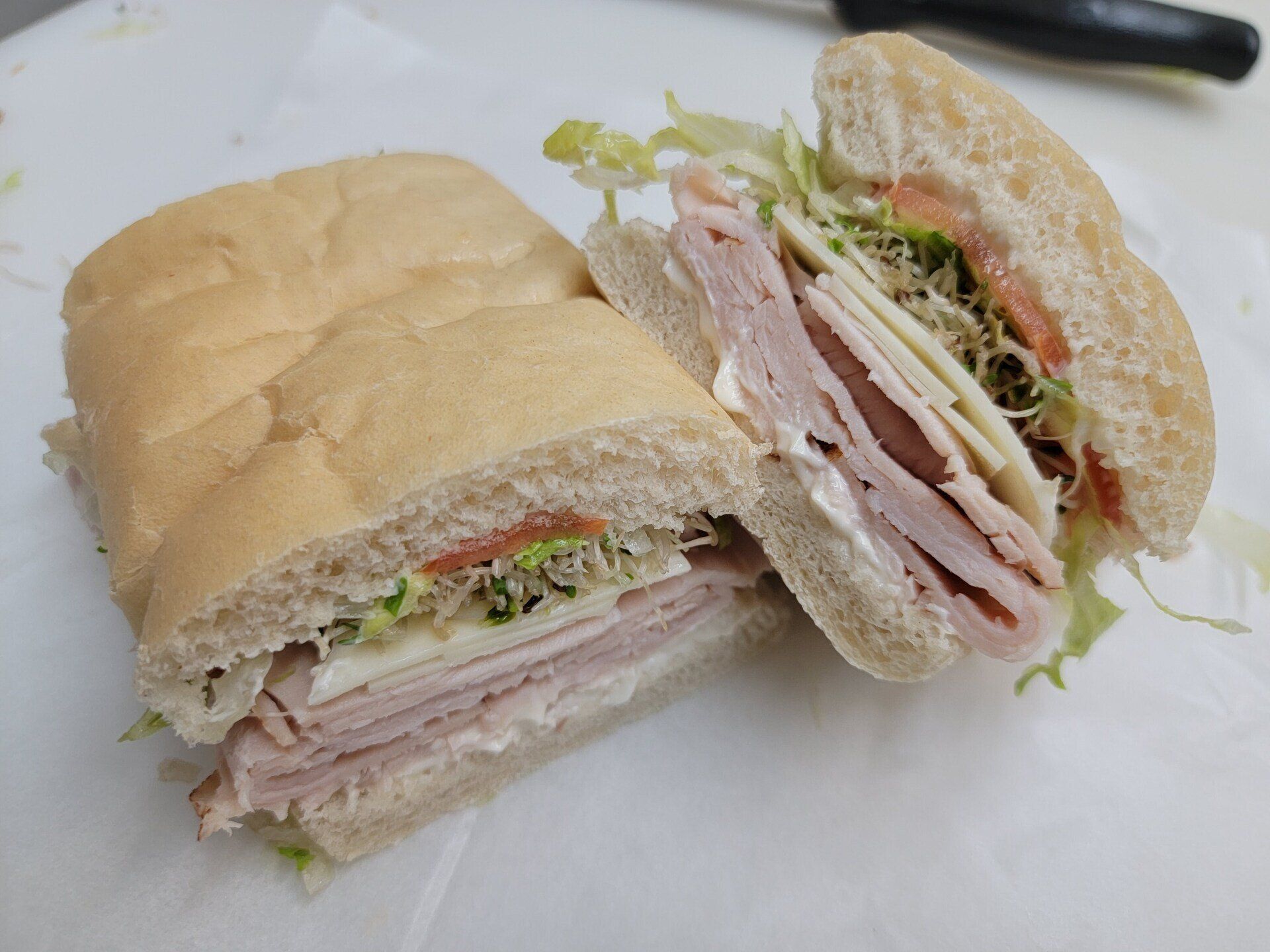 A halved ham and cheese sandwich on a crusty roll with lettuce and sprouts, sitting on white parchment paper.