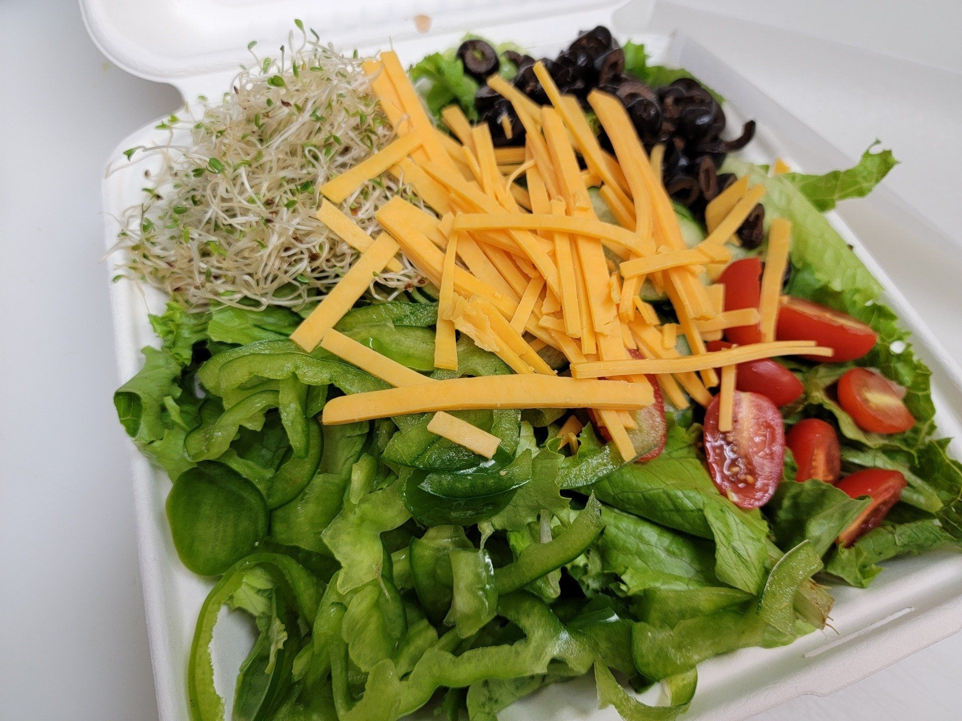 A green salad in a white takeout container topped with alfalfa sprouts, shredded cheddar cheese, olives, and tomatoes.