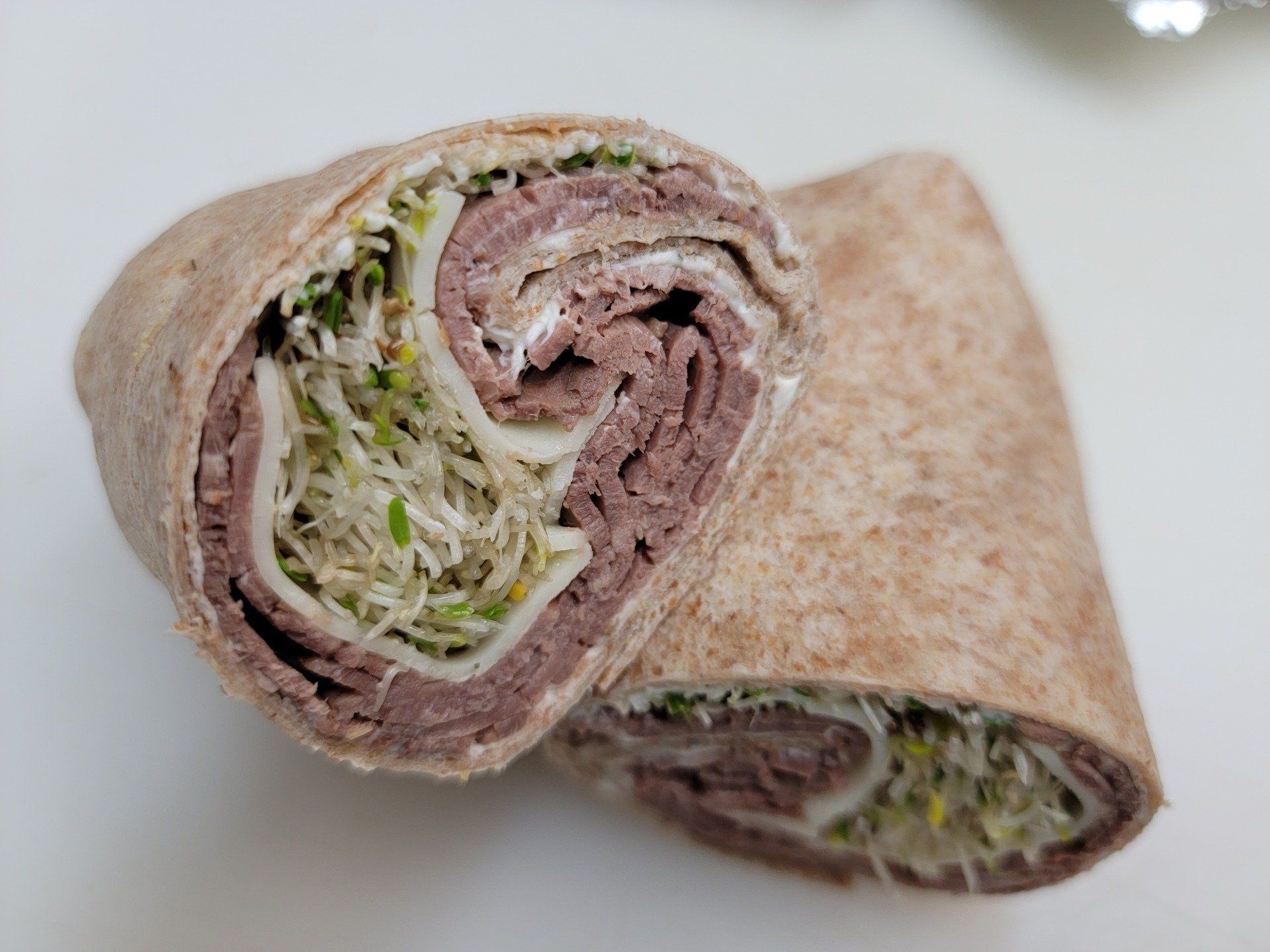 Two halves of a whole-wheat wrap filled with sliced roast beef, white cheese, and green sprouts on a white surface.