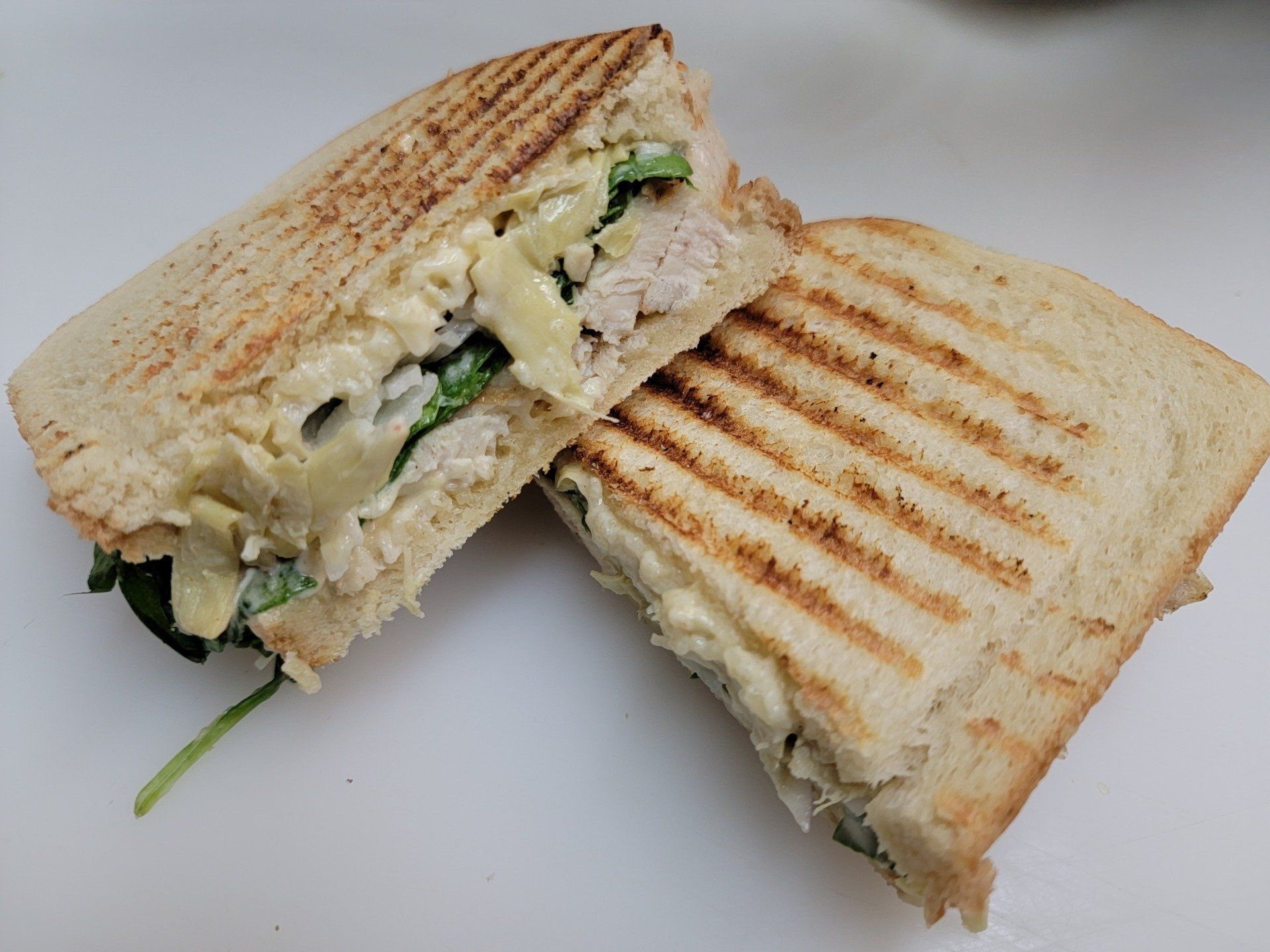 Two halves of a grilled panini sandwich on toasted bread with visible grill marks, filled with chicken, cheese, and greens.