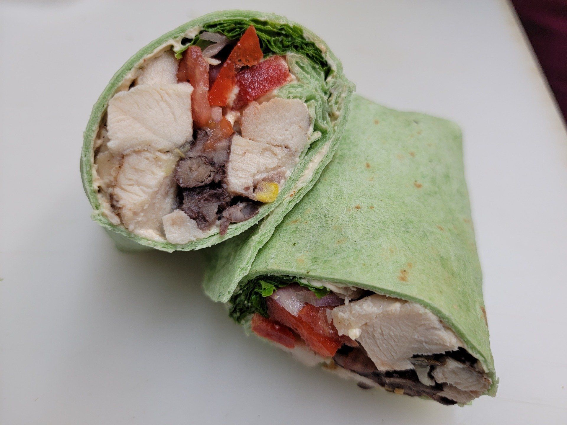 A green spinach tortilla wrap cut in half, revealing chicken, black beans, tomatoes, and greens on a white surface.