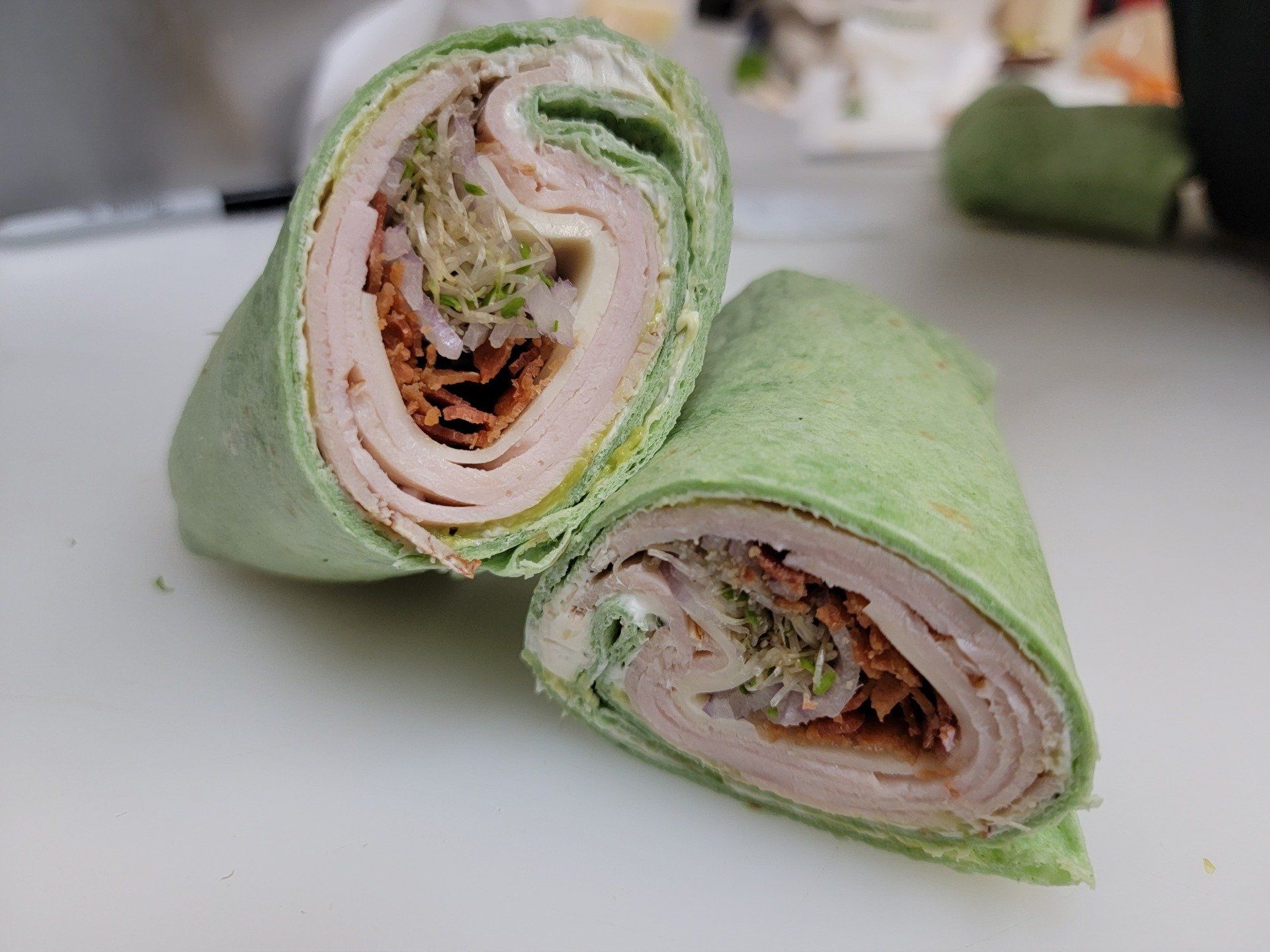 Two halves of a green spinach tortilla wrap filled with turkey, bacon, cheese, and sprouts on a white cutting board.