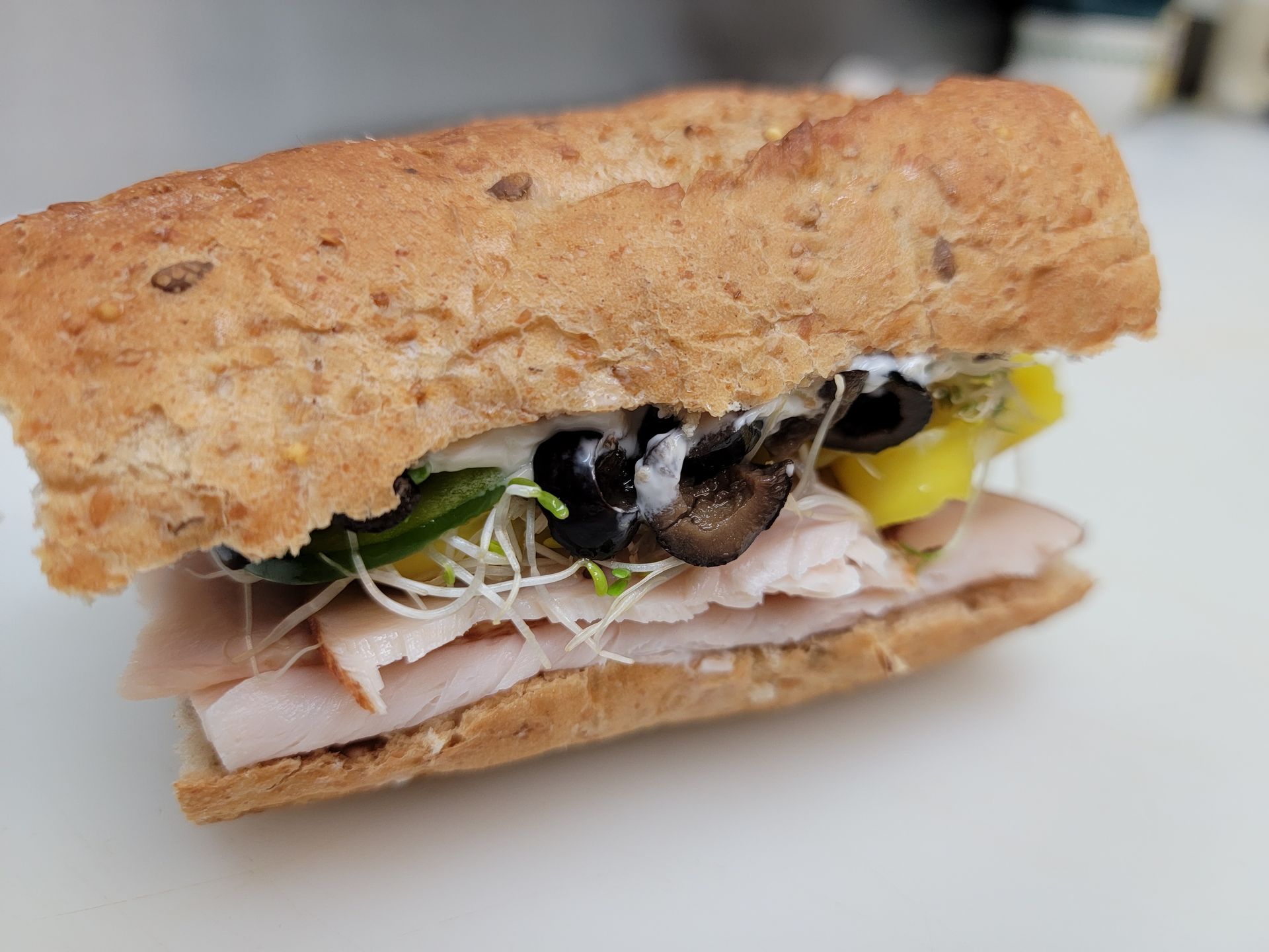 A sub sandwich on toasted wheat bread with turkey, sliced black olives, yellow peppers, sprouts, and white dressing.