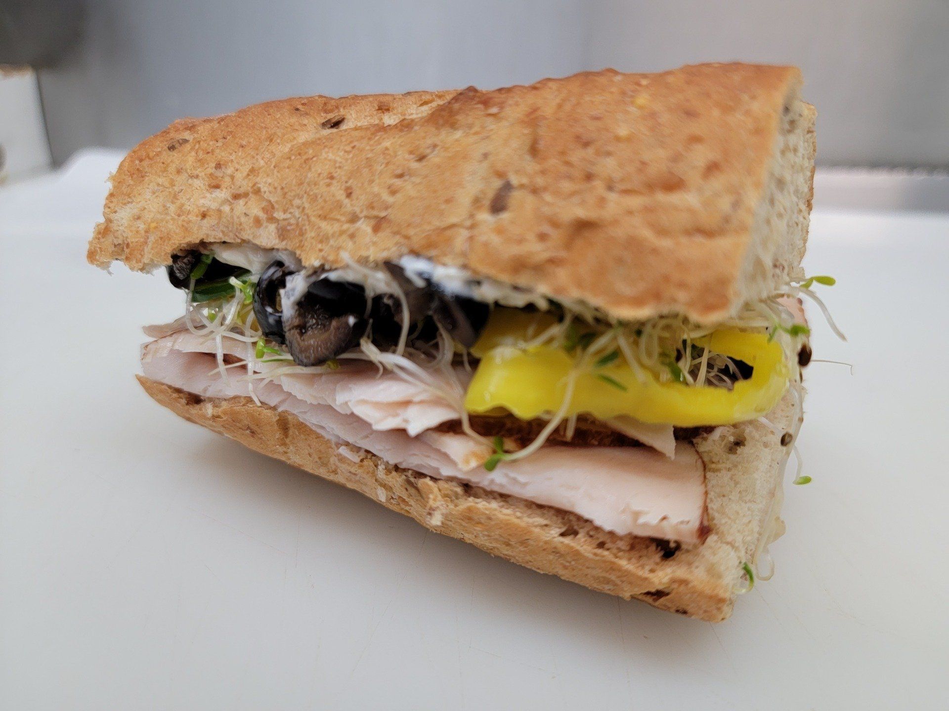 A turkey sandwich on a seeded baguette with olives, yellow pepper slices, sprouts, and dressing on a white background.