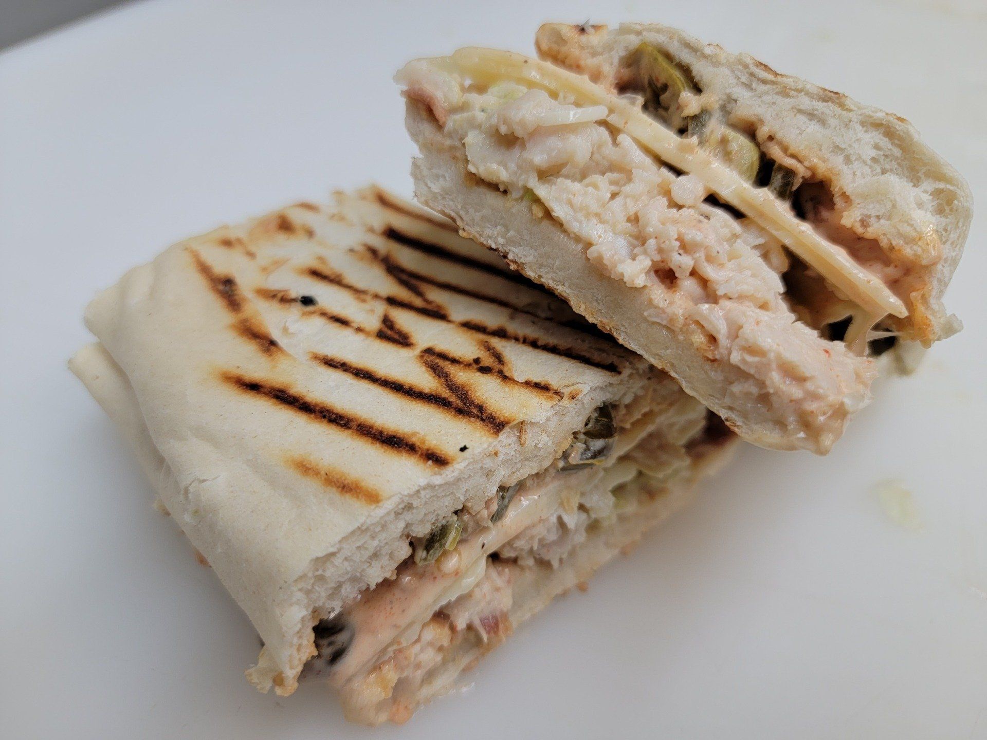 Two halves of a grilled panini with melted cheese, chicken or tuna salad, and sliced olives on a white surface.