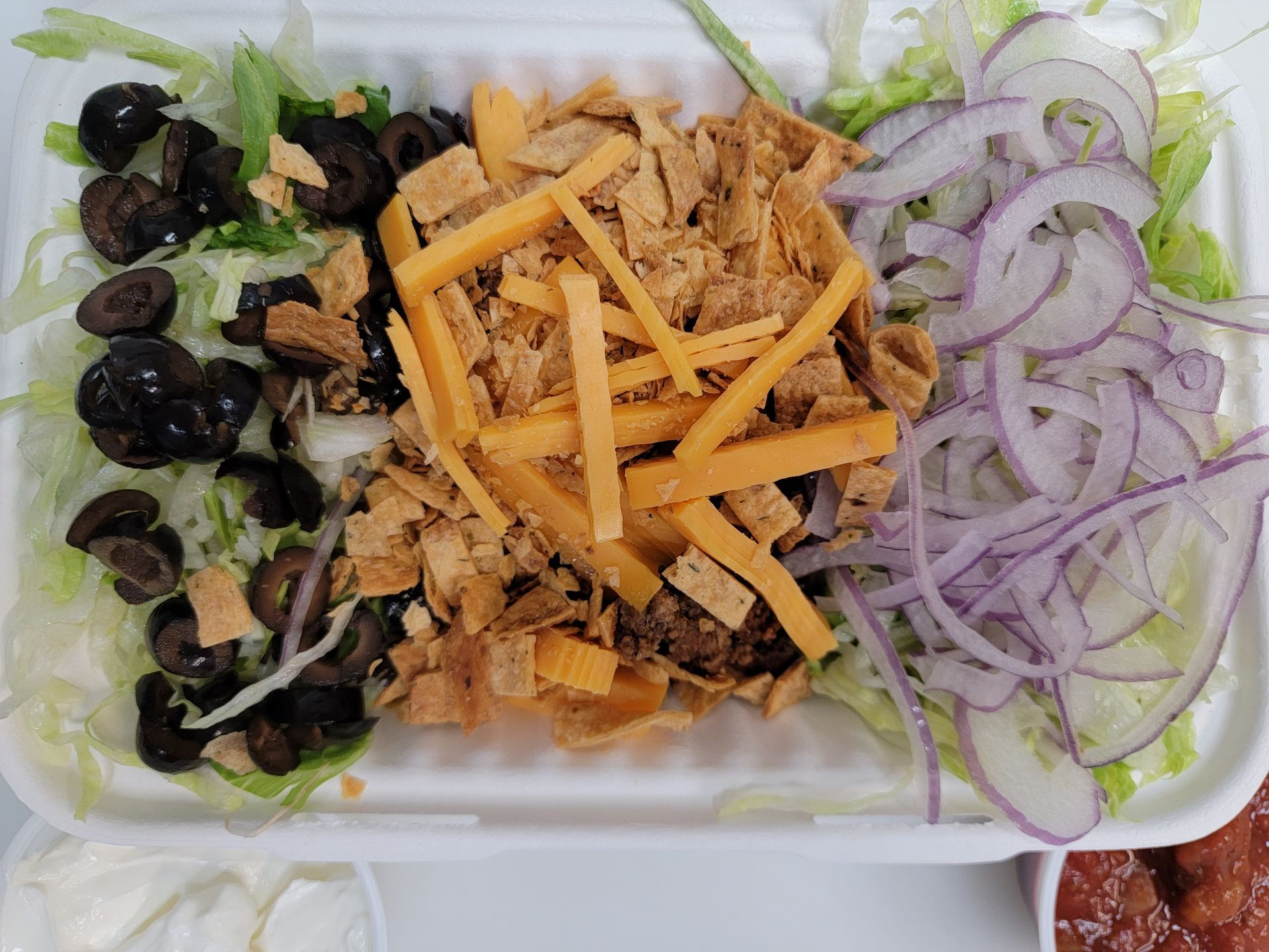 A rectangular takeout container with a salad topped with shredded cheddar, tortilla chips, black olives, and red onions.