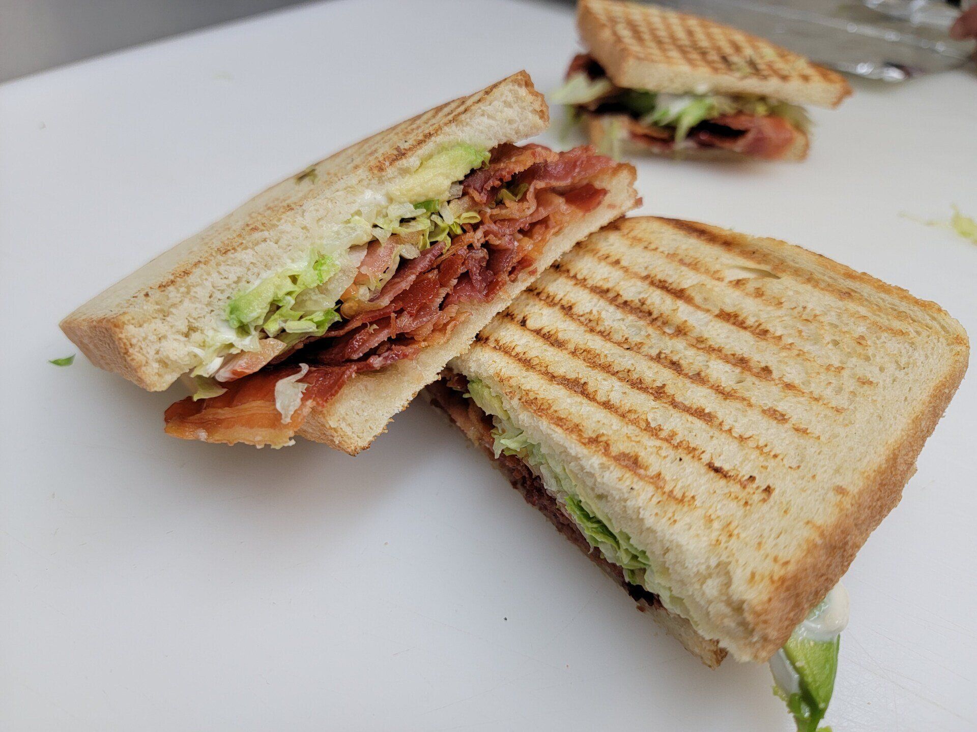 Grilled panini halves with bacon, lettuce, and avocado on toasted, striped bread sitting on a white surface.