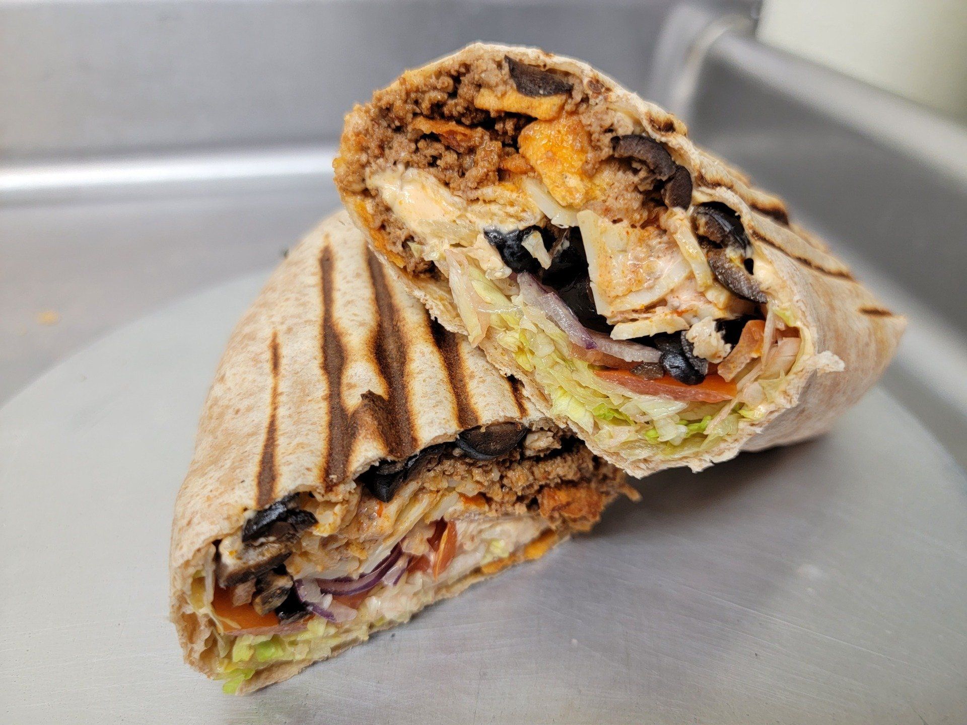 A cut, grilled wrap with ground meat, black olives, shredded lettuce, and sliced tomatoes on a silver tray.