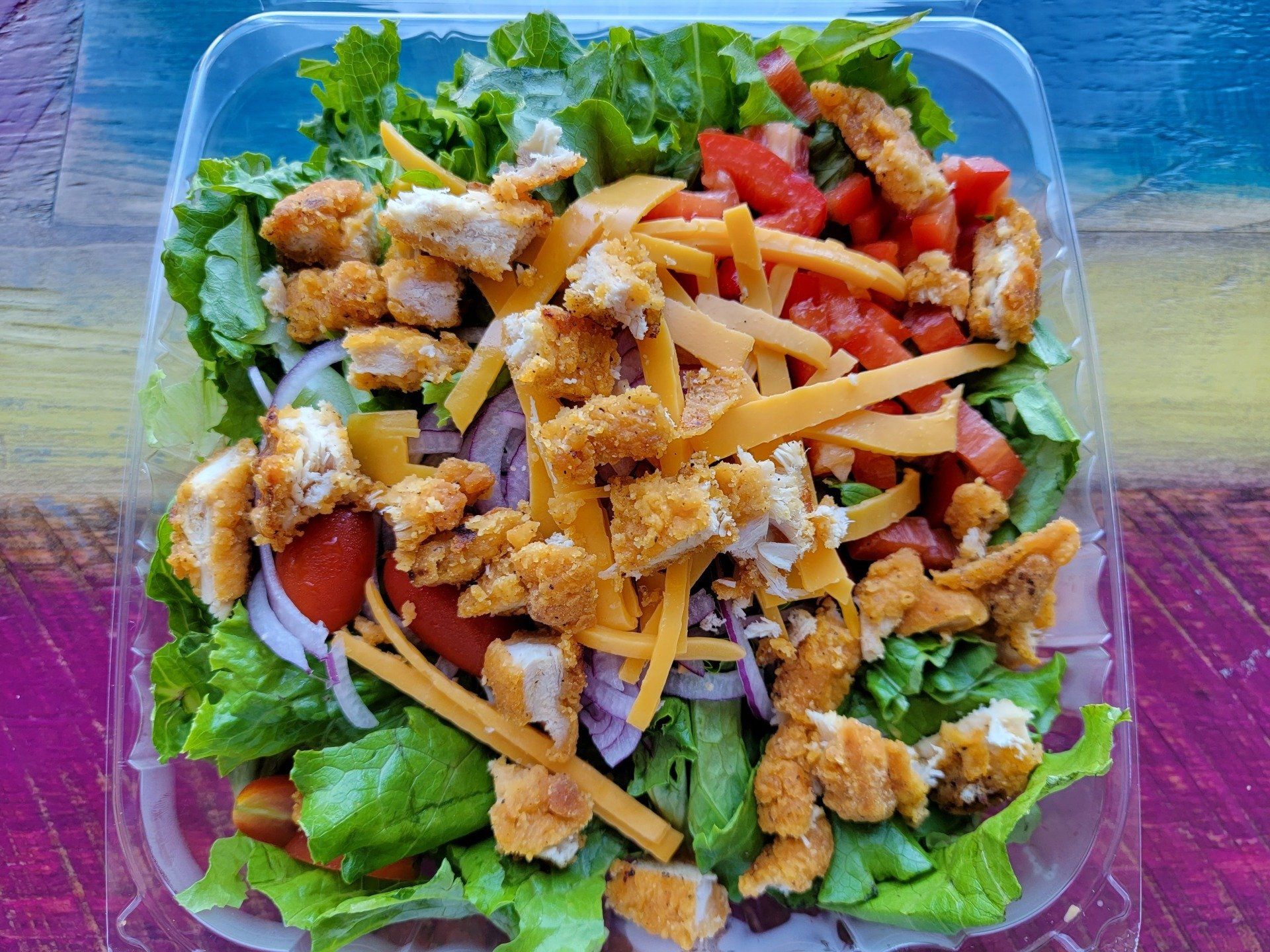 A salad in a clear plastic container featuring crispy chicken, shredded cheddar cheese, red tomatoes, and red onions.