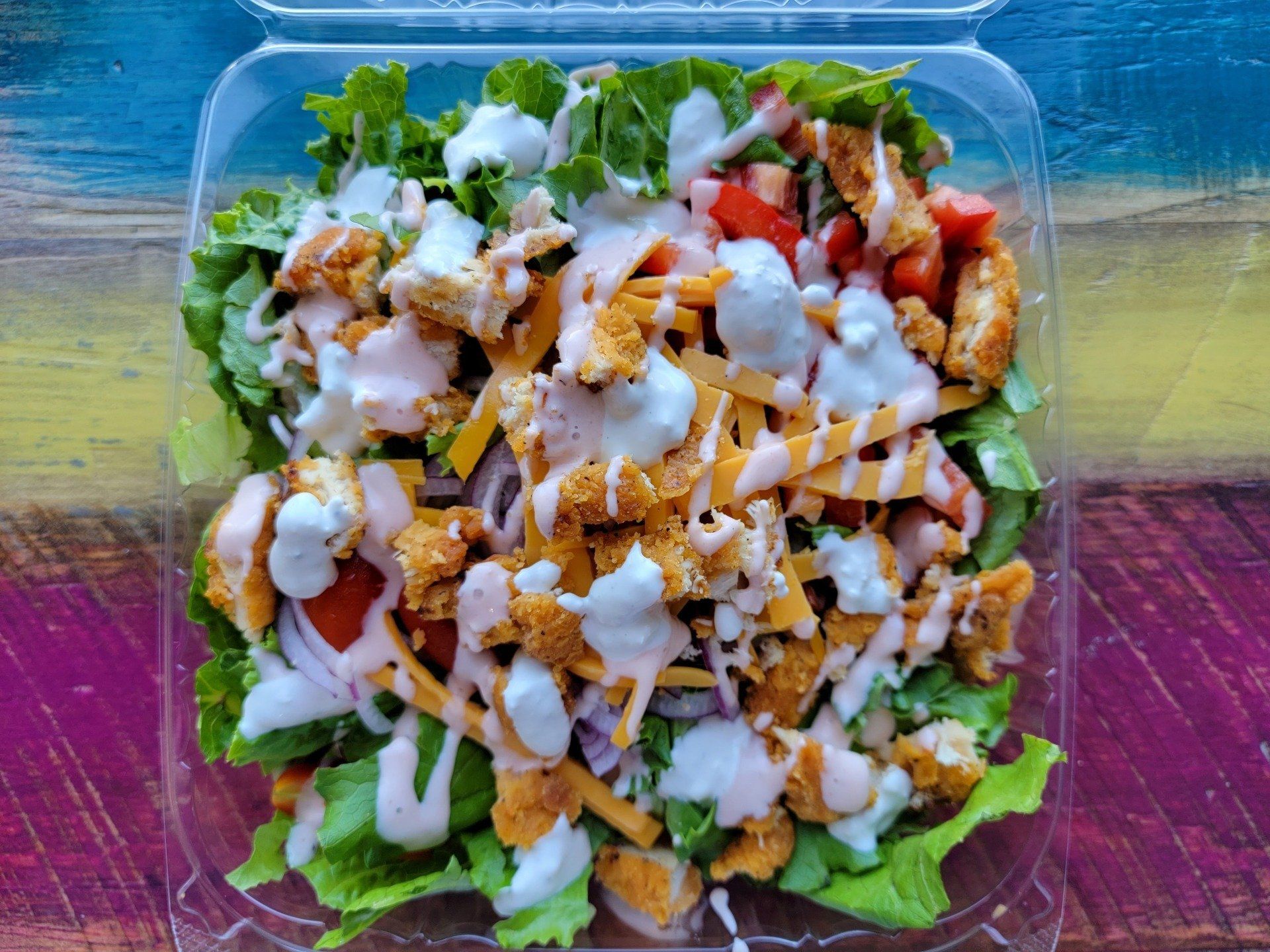 A clear plastic container filled with a crisp green salad topped with breaded chicken, cheddar cheese, and dressing.