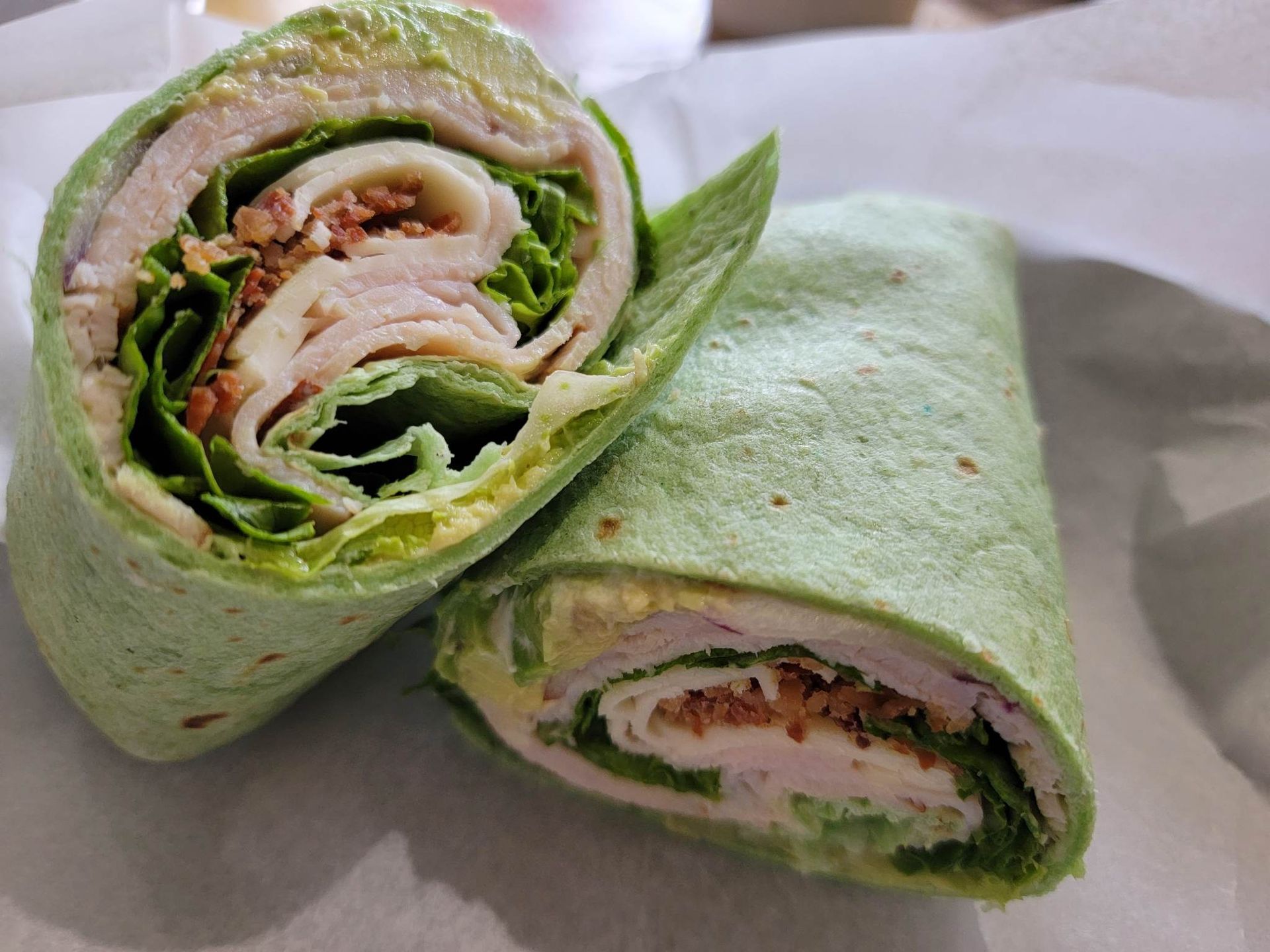 Two halves of a green spinach tortilla wrap filled with sliced turkey, lettuce, and bacon on parchment paper.