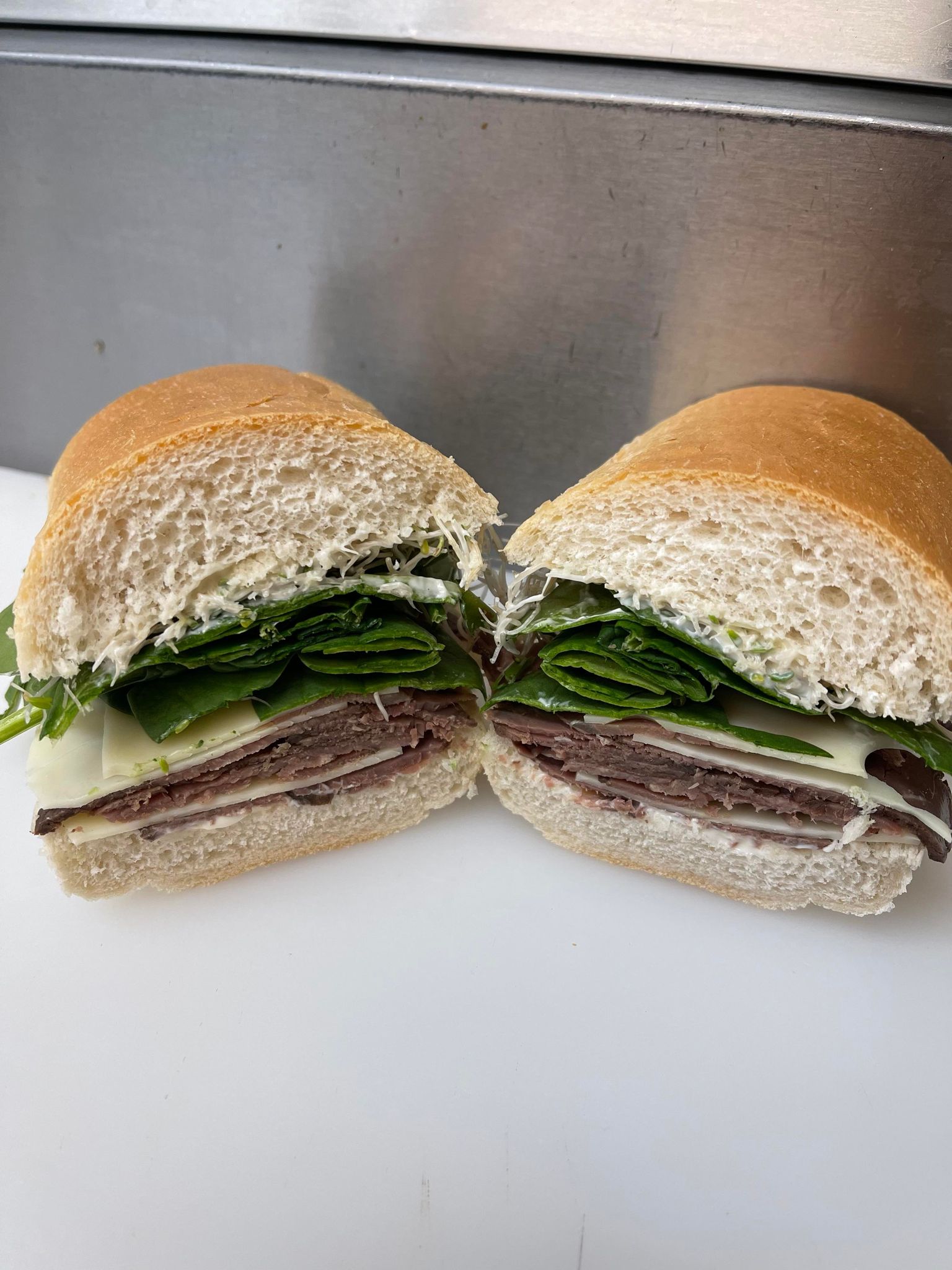A halved sandwich on a white surface, featuring sliced meat, white cheese, and dark leafy greens on a light bread roll.