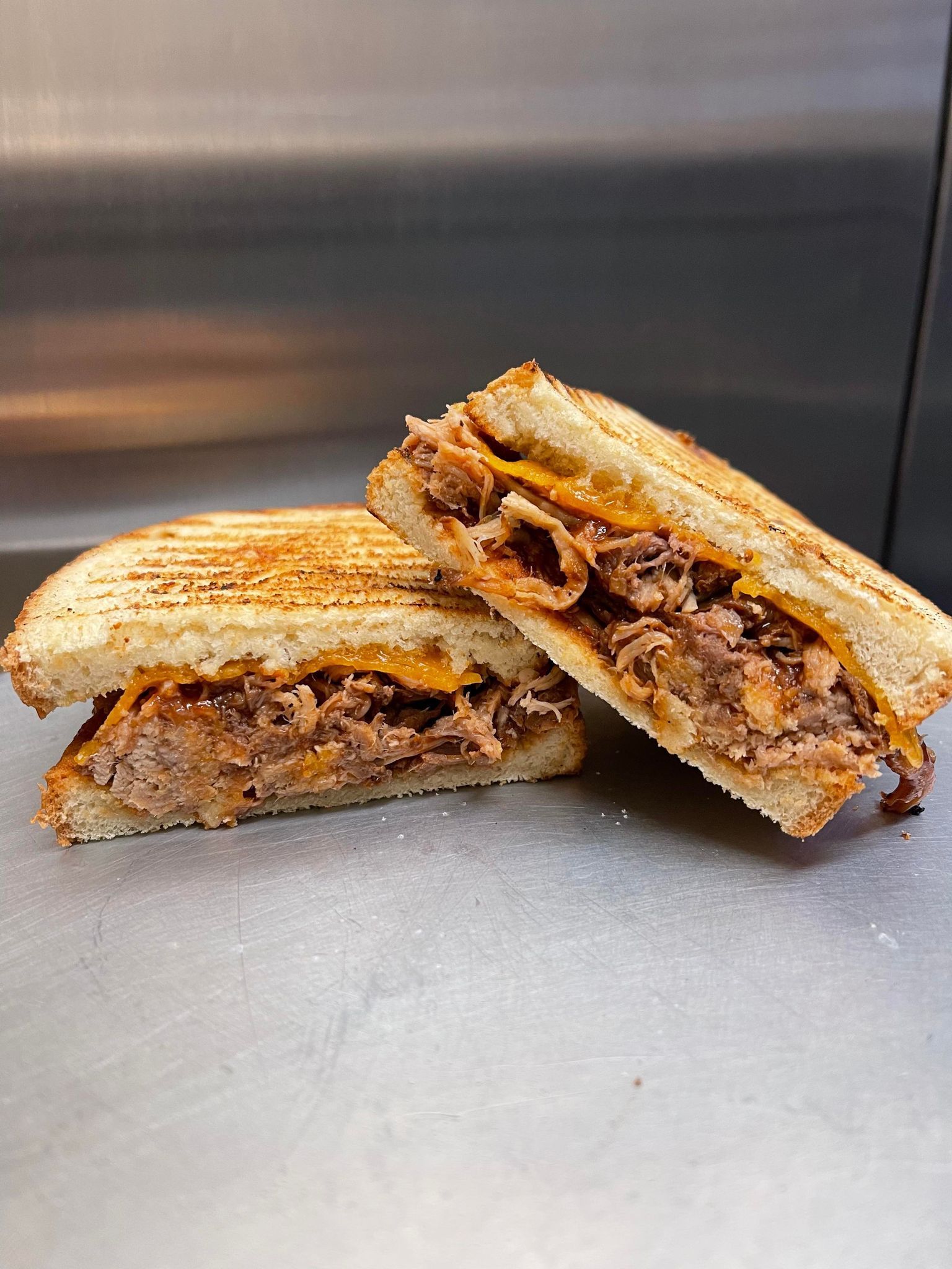 Two halves of a grilled, golden-brown pulled pork and melted cheese sandwich sitting on a metallic surface.