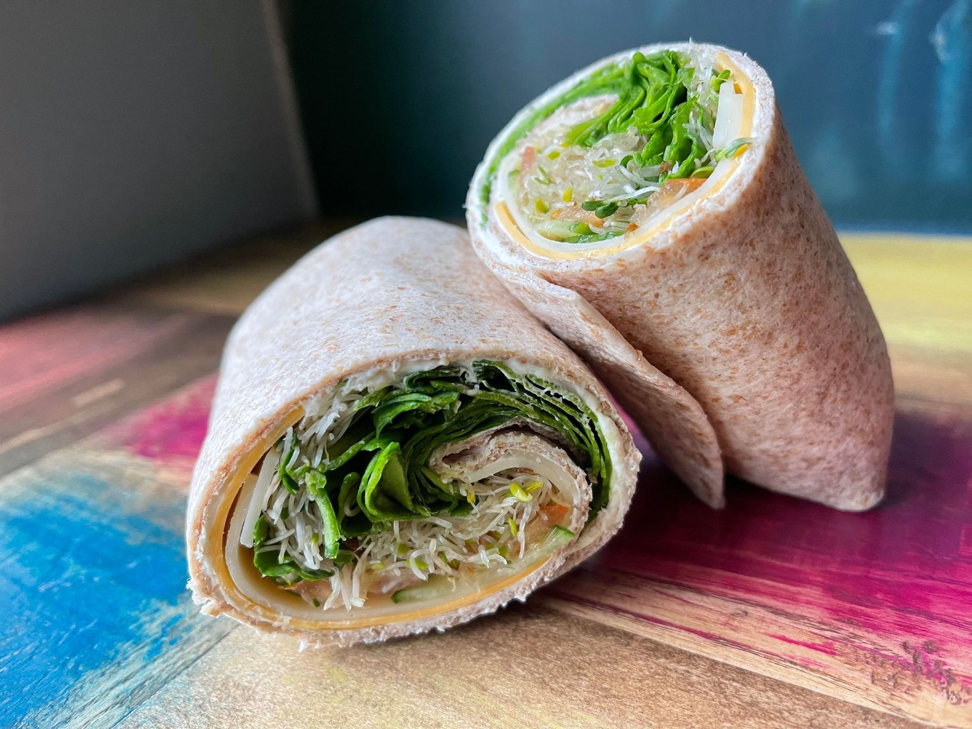 Two halves of a whole-wheat wrap filled with cheese, sprouts, and greens, resting on a colorful, painted wooden surface.