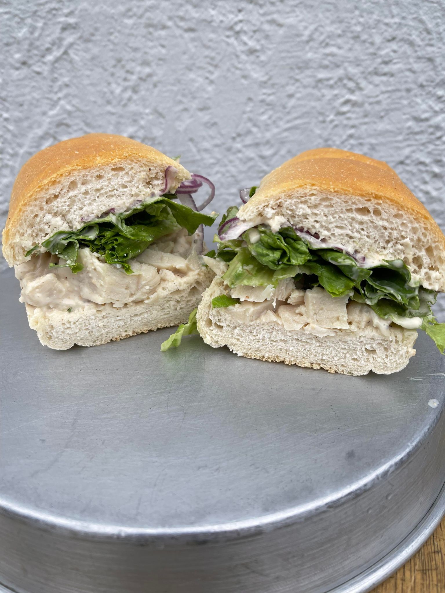 Two halves of a chicken salad sandwich on a round metal tray against a textured wall.