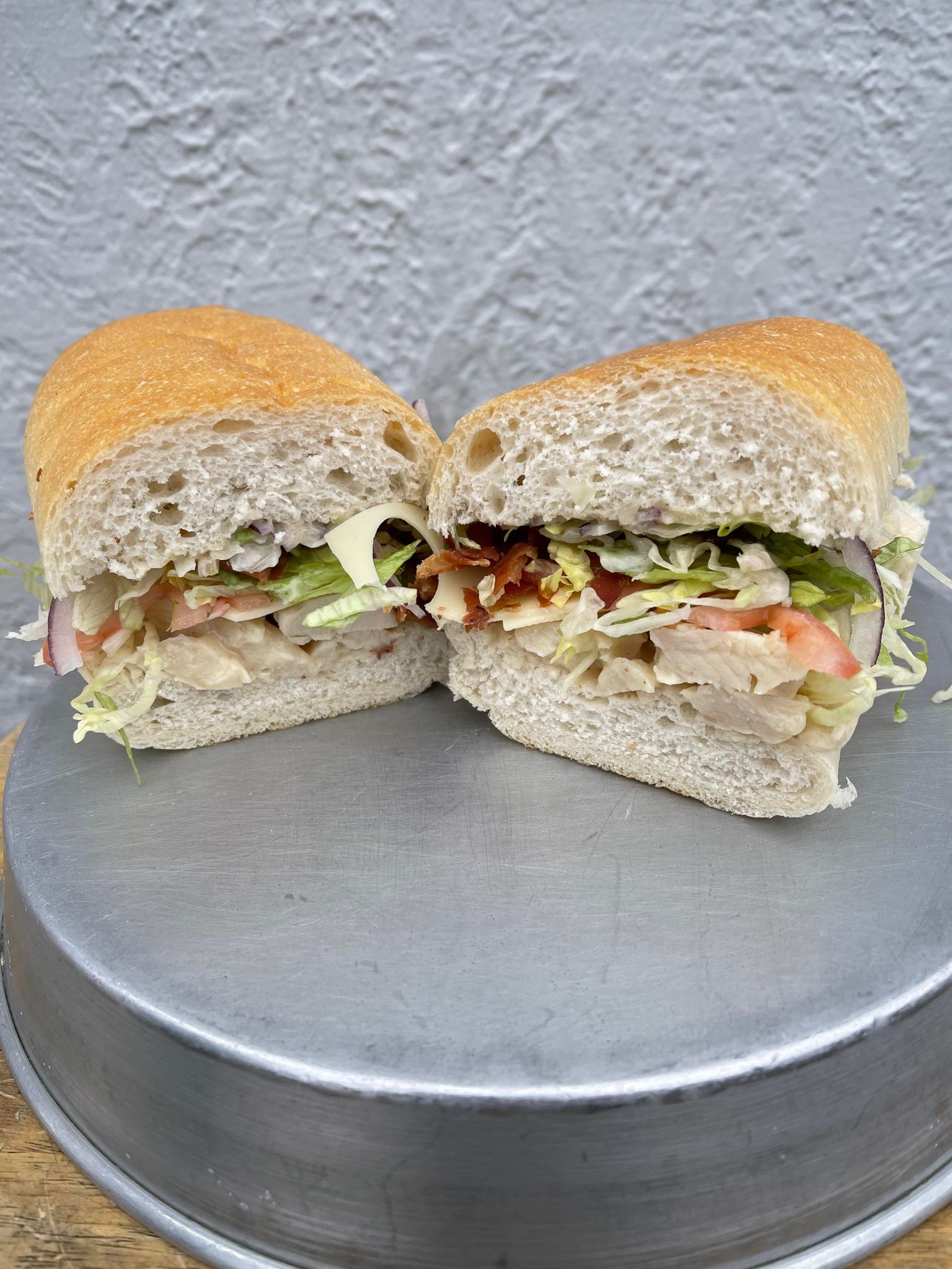 A chicken sandwich cut in half on a hoagie roll, filled with lettuce, tomato, and bacon, resting on a metal tray.