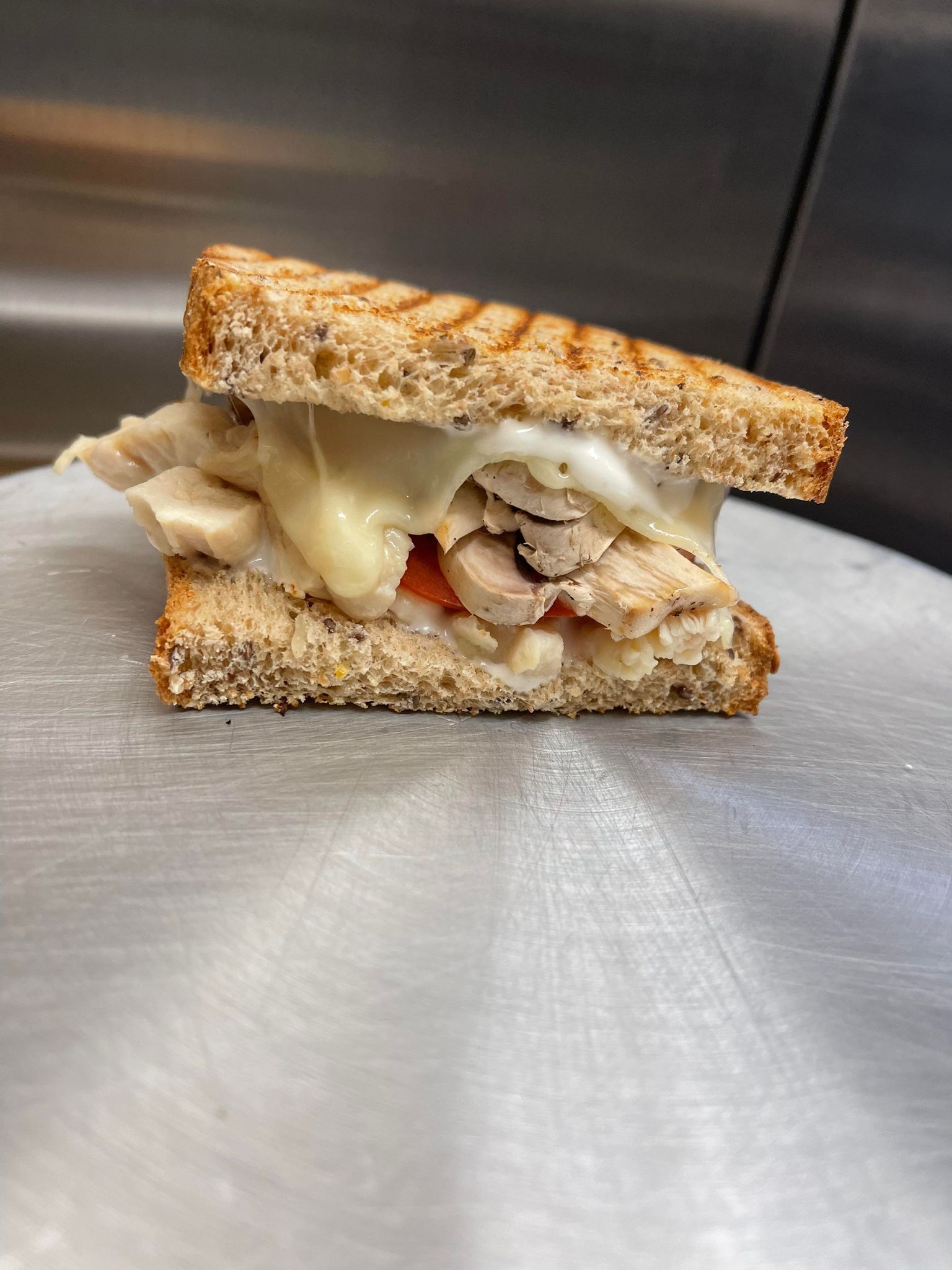 A grilled chicken and melted cheese sandwich on whole-grain bread, sitting on a metallic surface.